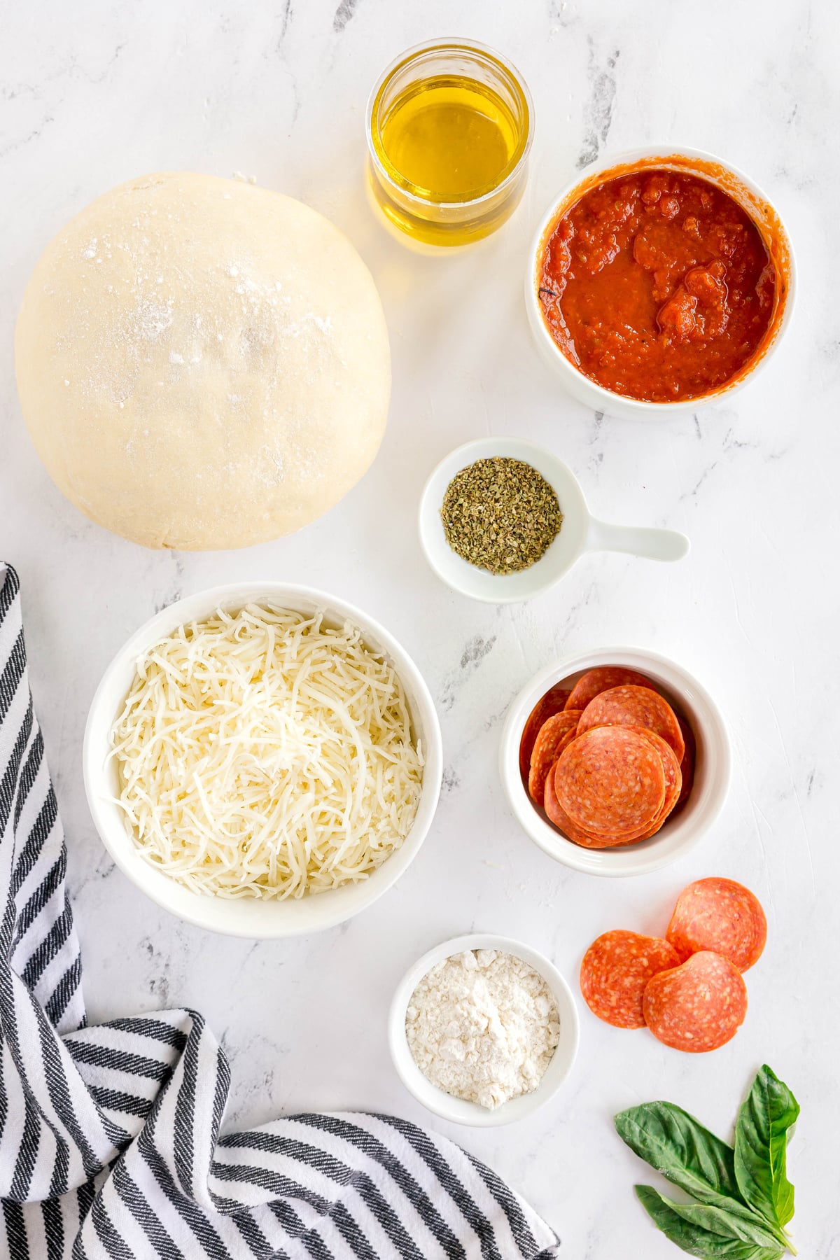 Homemade Pepperoni Pizza ingredients on a white surface: pizza dough, olive oil, pizza sauce, dried oregano or Italian seasoning, mozarella cheese, pepperoni, and flour with basil leaves and linen.