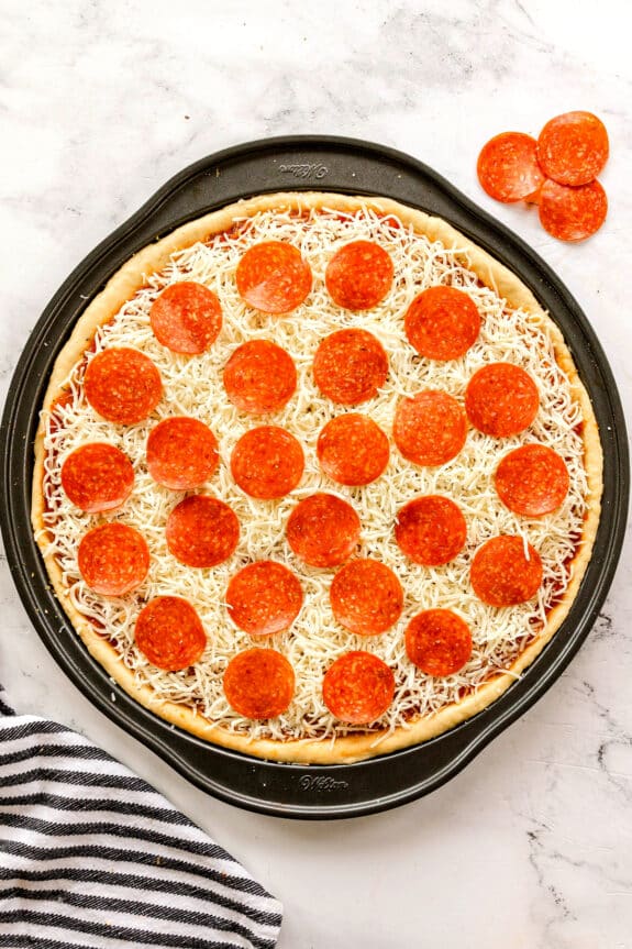 Unbaked Homemade Pepperoni Pizza with shredded cheese and pepperoni slices on a round baking pan, with extra pepperoni slices on the side.