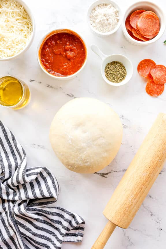 Pizza dough, flour, shredded cheese, tomato sauce, pepperoni slices, dried herbs, olive oil, a striped towel, and a rolling pin for Homemade Pepperoni Pizza on a white marble surface.