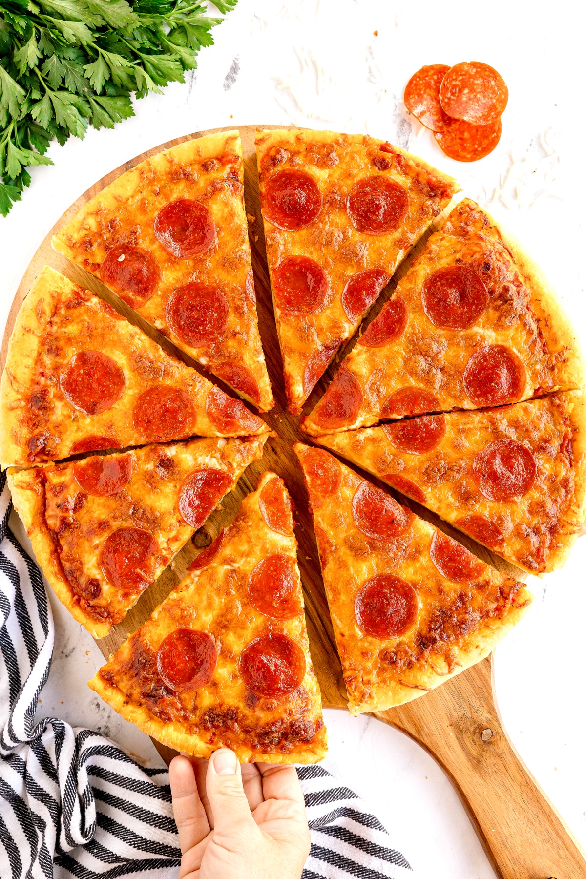 A Homemade Pepperoni Pizza cut into eight slices on a wooden paddle, with one slice being lifted by a hand. Parsley and pepperoni slices are in the background.