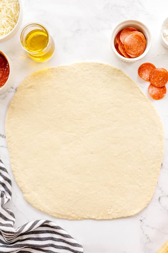 A rolled-out pizza dough for Homemade Pepperoni Pizza recipe on a marble surface, surrounded by bowls of shredded cheese, olive oil, pizza sauce, pepperoni slices, and a striped towel.