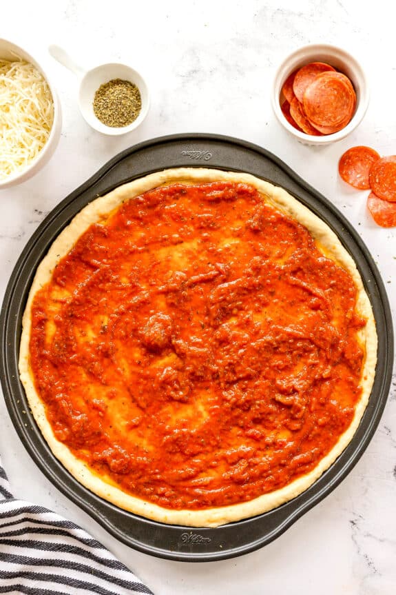 Unbaked Homemade Pepperoni Pizza crust on a pan topped with tomato sauce, surrounded by bowls of shredded cheese, dried herbs, and pepperoni slices.