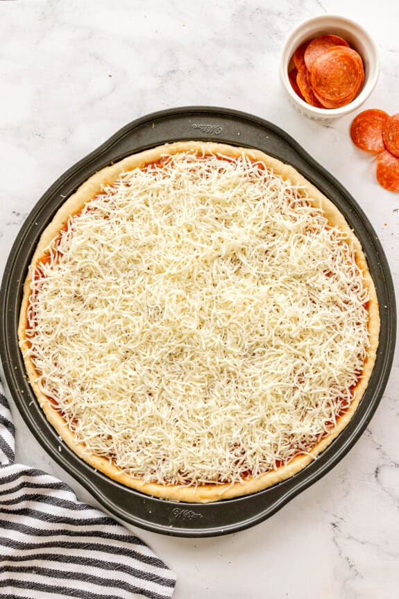Unbaked Homemade Pepperoni Pizza with shredded cheese on dough in a round baking pan, next to a bowl and slices of pepperoni on a marble surface.