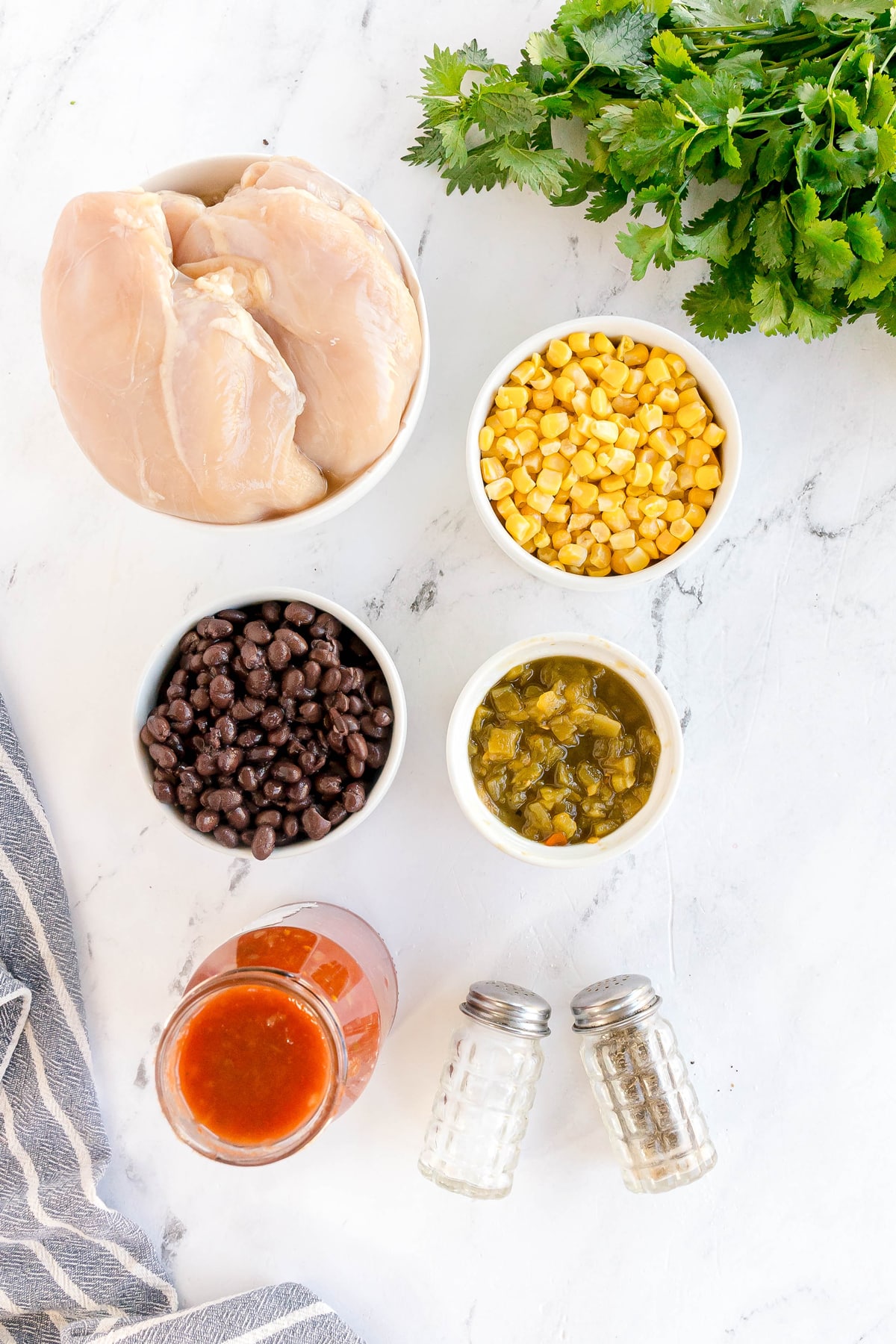 Overhead view of ingredients for Slow Cooker Southwest Chicken: raw skinless boneless chicken breasts, black beans, corn, green chiles, salsa, salt and pepper, cilantro, and a striped towel on a white countertop.