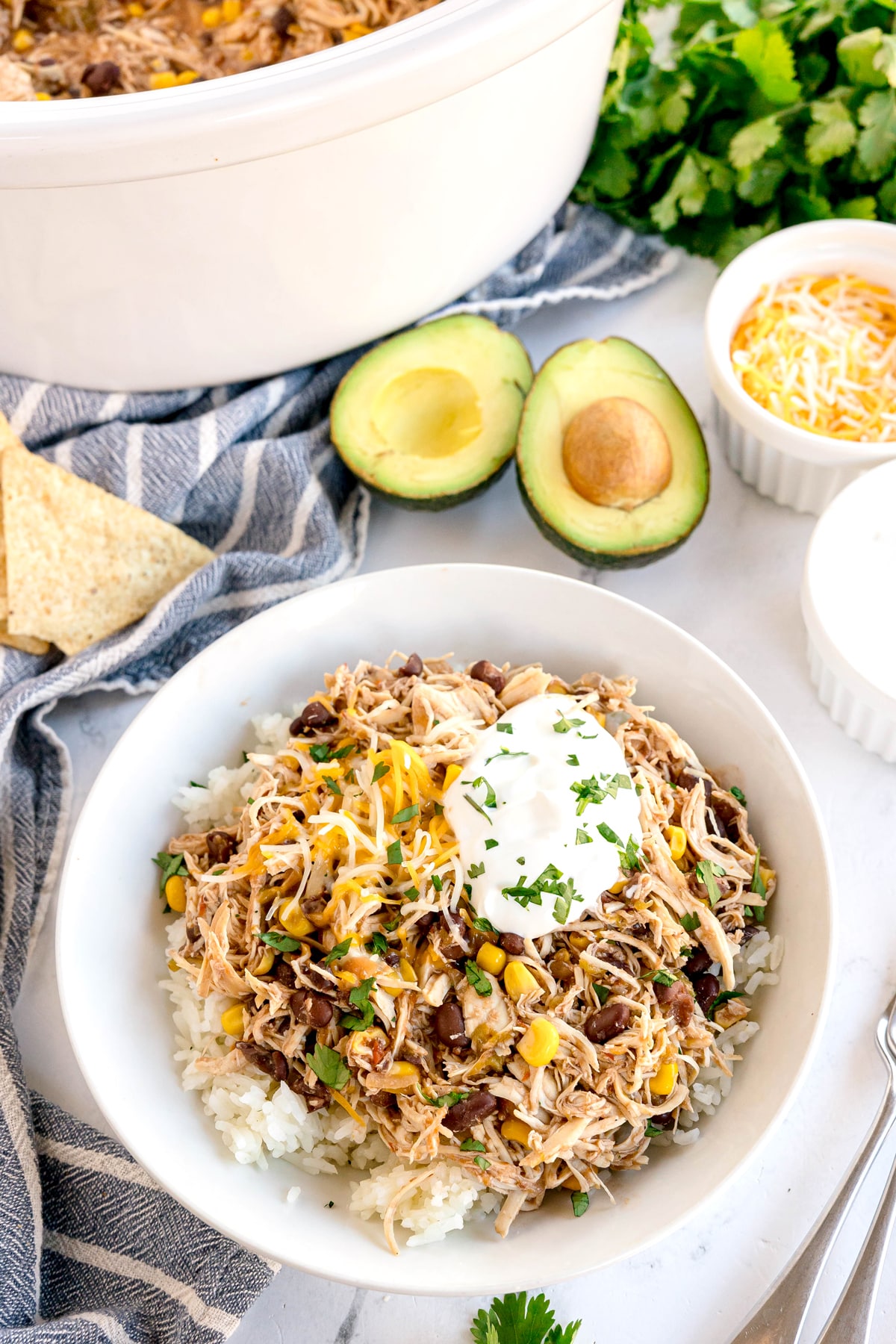 A bowl of Slow Cooker Southwest Chicken with shredded chicken, beans, corn, rice, shredded cheese, and sour cream, garnished with parsley, sits beside avocado halves and small bowls of toppings.