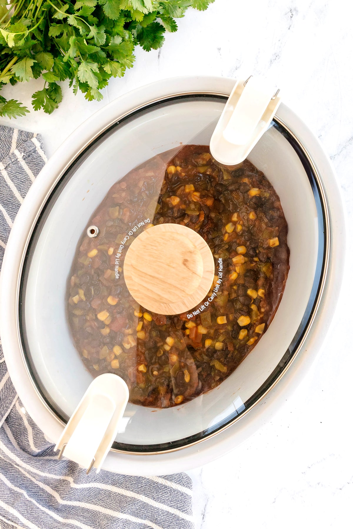 A slow cooker with a glass lid contains a mixture of beans, corn, and vegetables in a dark sauce. Fresh cilantro and a striped towel are beside it. This is one process in preparing Slow Cooker Southwest Chicken