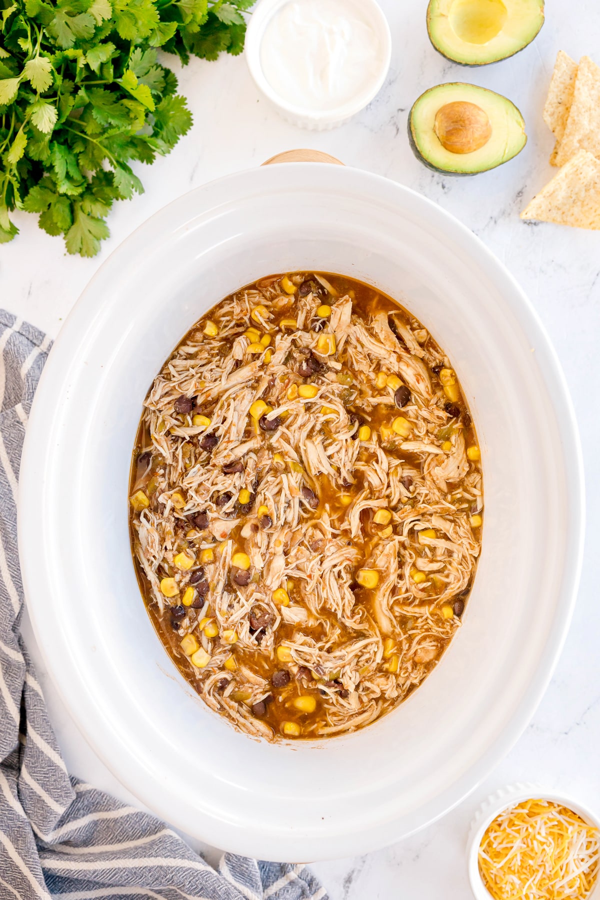 Slow Cooker Southwest Chicken with shredded chicken, corn, and black beans in broth in a white slow cooker, surrounded by cilantro, avocado, tortilla chips, cheese, and sour cream.