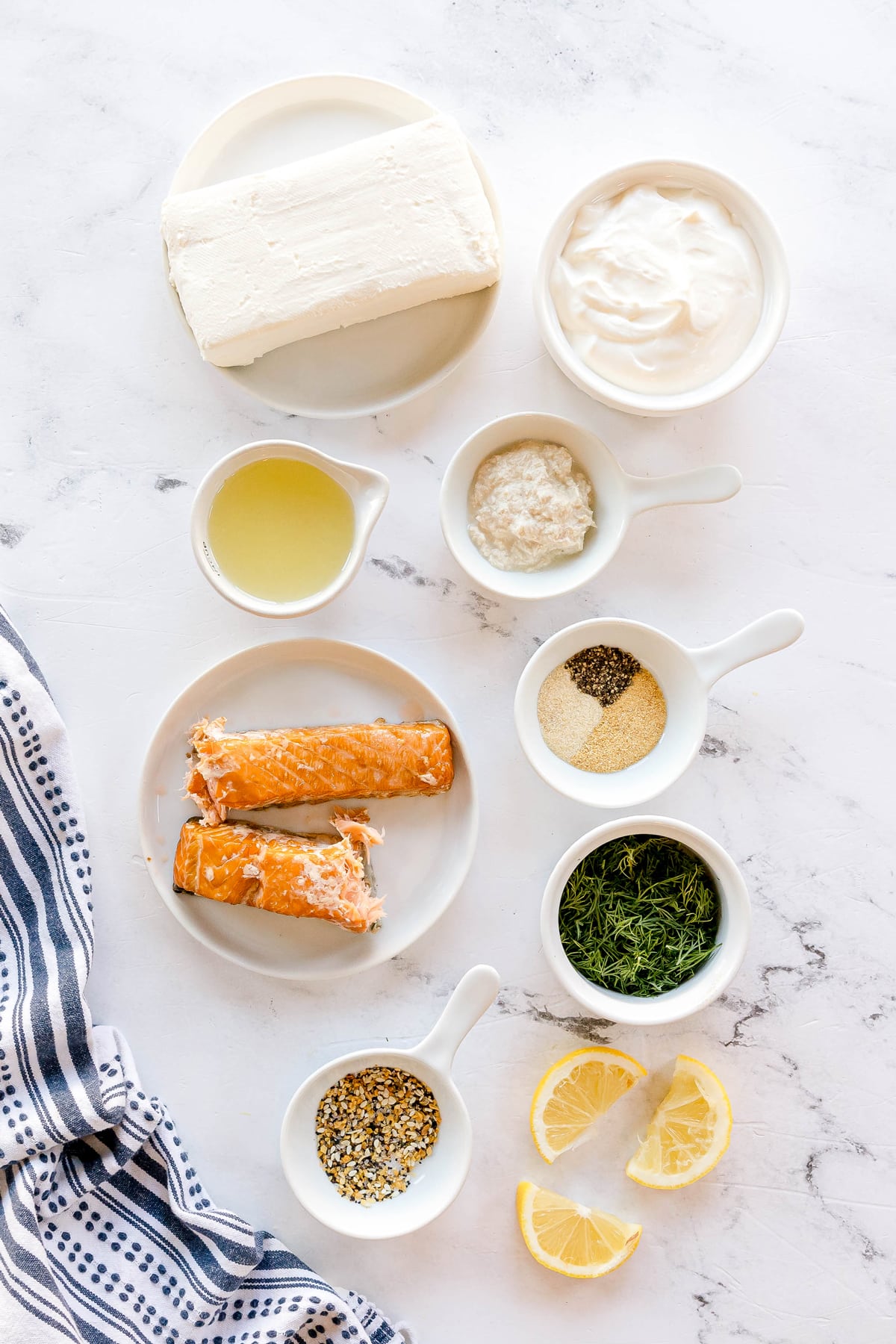 Overhead view of Smoked Salmon Dip Recipe ingredients on white surface: cream cheese, sour cream, lemon juice, horseradish, finely-chopped smoked salmon, garlic, onion powder, black pepper, chopped dill, bagel seasoning, lemon wedges, assorted crackers and vegetables, and a striped towel.