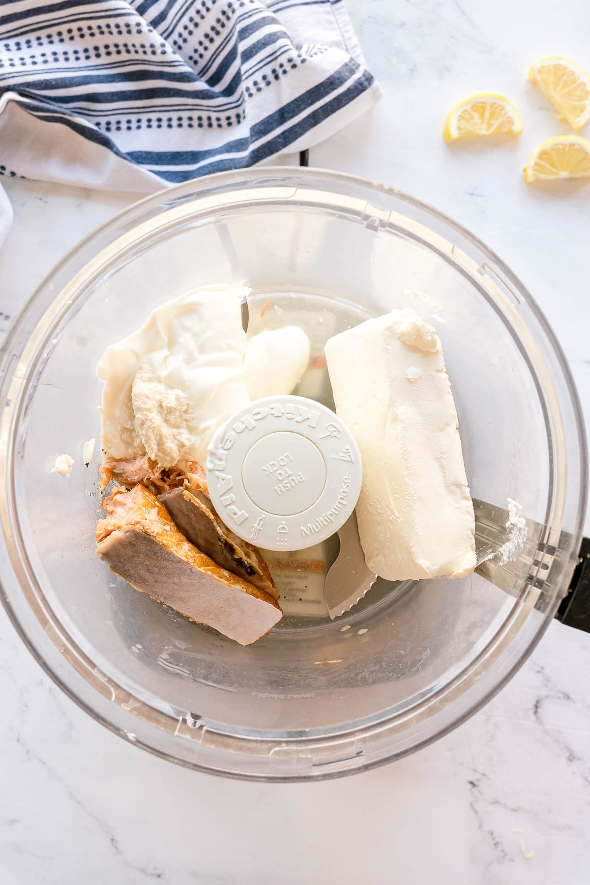 A food processor bowl containing cream cheese, sour cream, lemon juice, horseradish, garlic powder, onion powder, black pepper, and smoked salmon with lemon slices and a striped cloth nearby. This is the first step in preparing Smoked Salmon Dip Recipe.