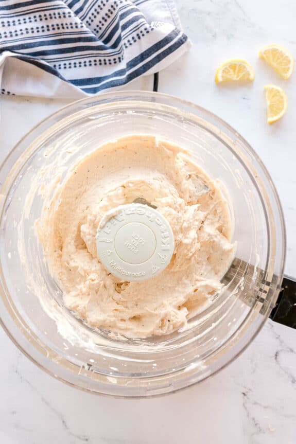 Smoked Salmon Dip Recipe in a glass mixing bowl with creamy batter is set on a marble countertop, next to a striped towel and three lemon wedges.