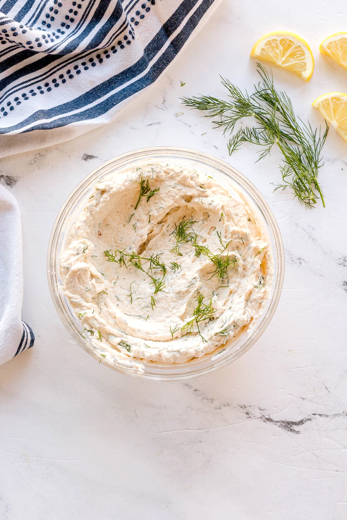A glass bowl of Smoked Salmon Dip Recipe garnished with fresh dill sits on a marble surface, with lemon wedges and dill sprigs nearby and a striped cloth in the corner.