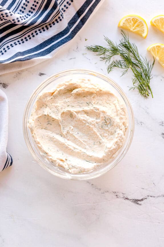 A glass bowl filled with Smoked Salmon Dip Recipe sits on a marble surface near lemon wedges, fresh dill, and a striped towel.
