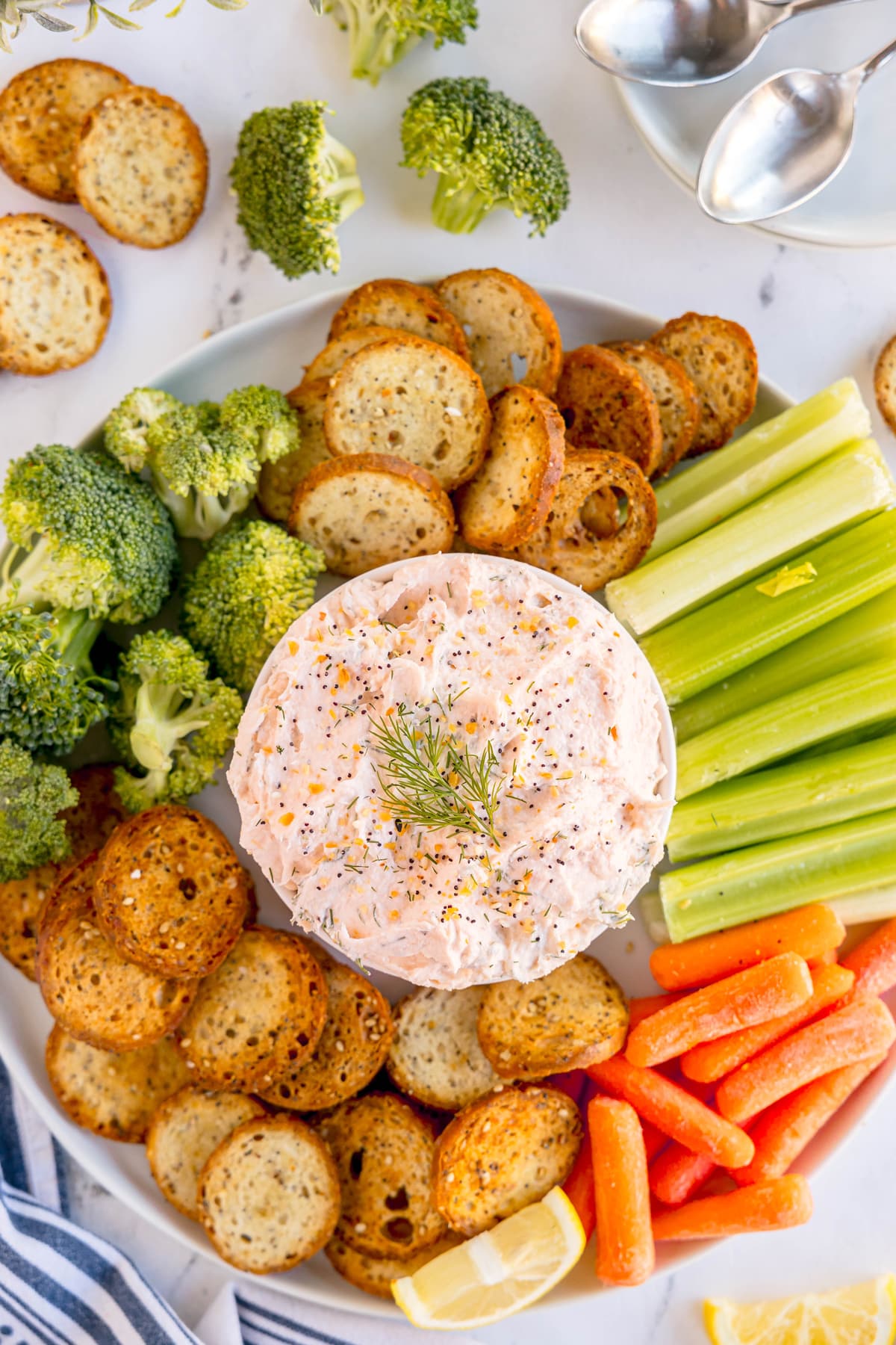 A plate with a bowl of Smoked Salmon Dip Recipe with dill, surrounded by bagel chips, celery sticks, baby carrots, and broccoli florets.