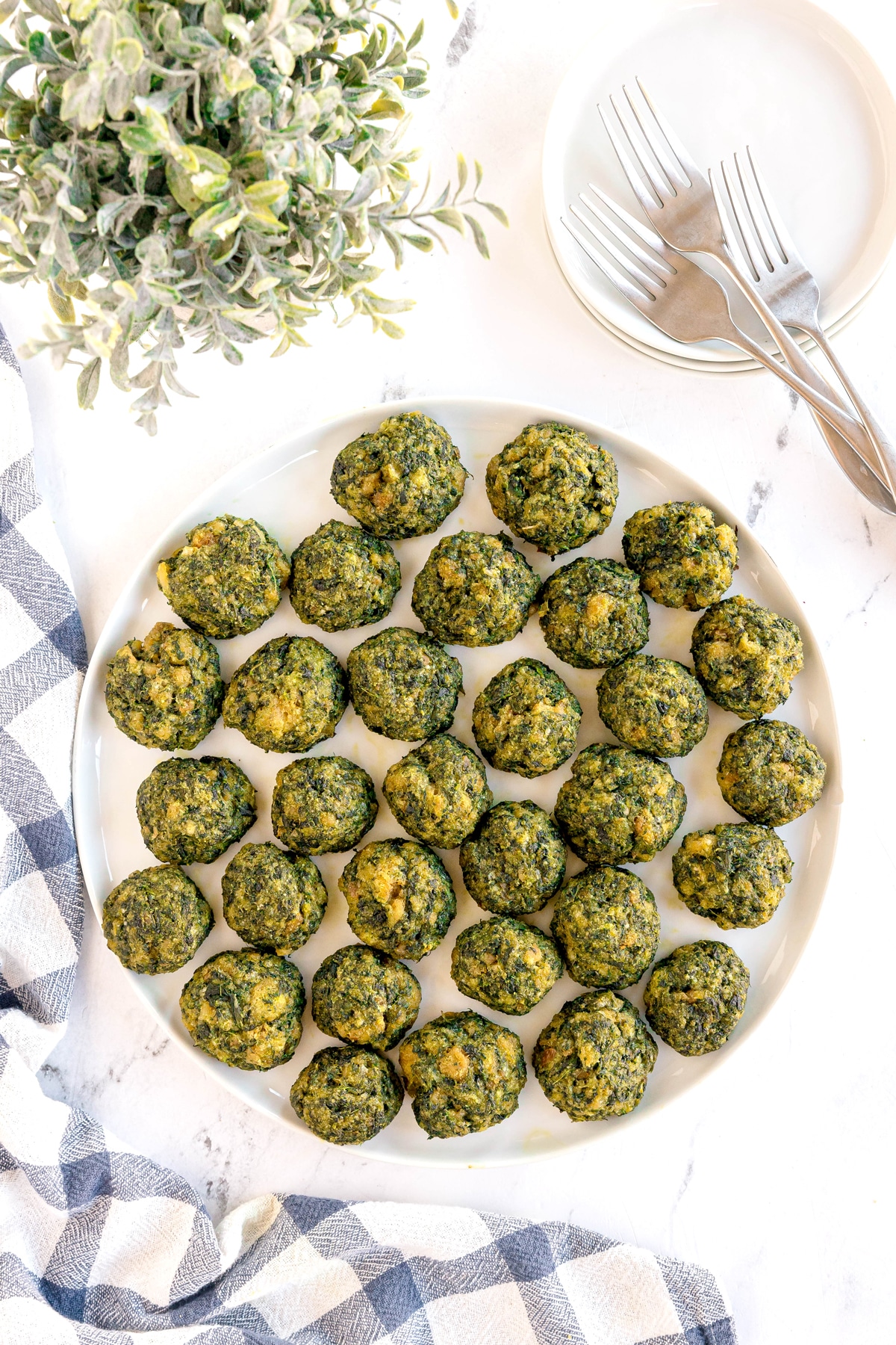 A white plate with evenly spaced spinach balls on a marble surface, next to cutlery, stacked plates, a green plant, and a blue-and-white checkered cloth.