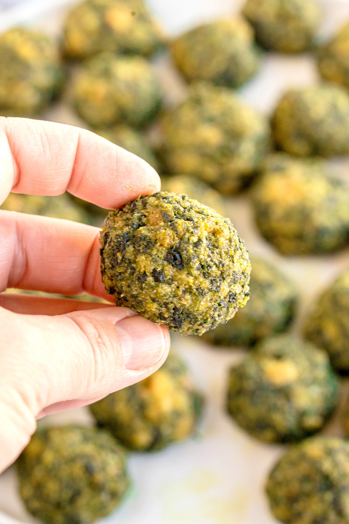 A hand holding a round, green and yellow baked spinach ball, with more spinach balls blurred in the background.