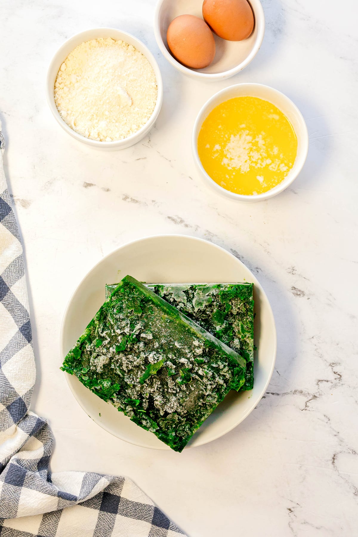 A plate with two blocks of frozen spinach, a bowl of flour, two eggs in a bowl, and a bowl of melted butter on a white countertop for Spinach Balls recipe.