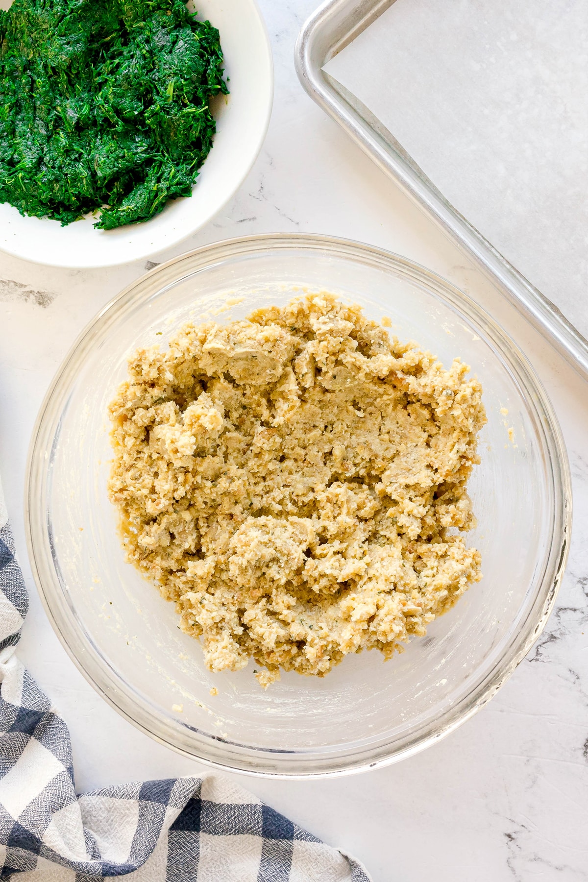 A glass bowl filled with a stuffing mixture sits on a marble surface next to a bowl of chopped spinach, a striped towel, and a baking sheet lined with parchment paper. These are for spinach balls recipe.