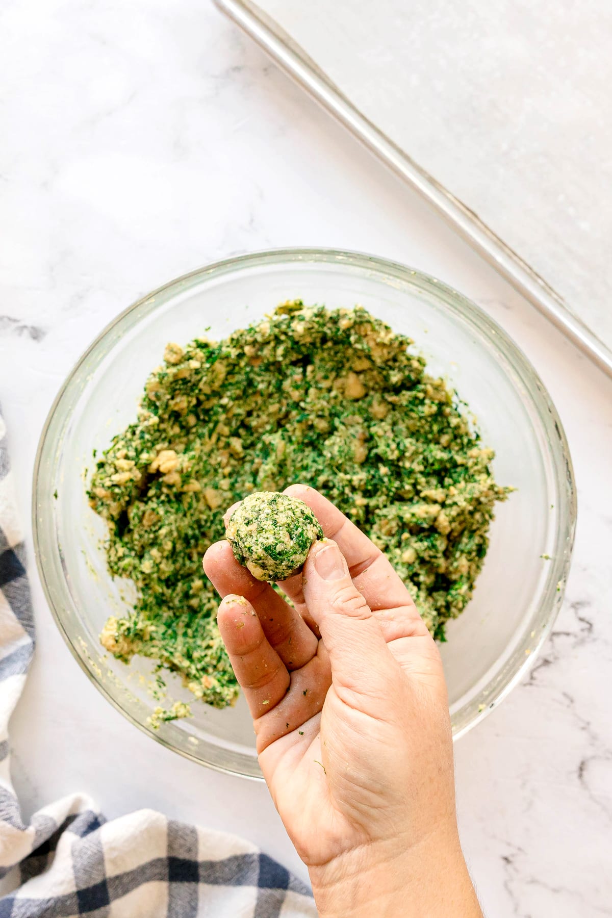A hand holds a round ball of uncooked Spinach Balls mixture above a glass bowl filled with more mixture on a white marble surface.