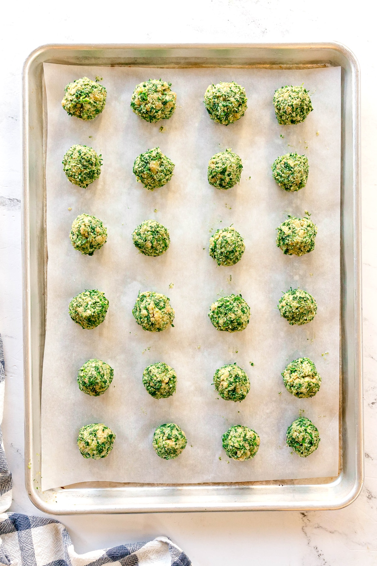 A baking sheet lined with parchment paper holds 24 evenly spaced, uncooked green and yellow vegetable balls arranged in a grid pattern.