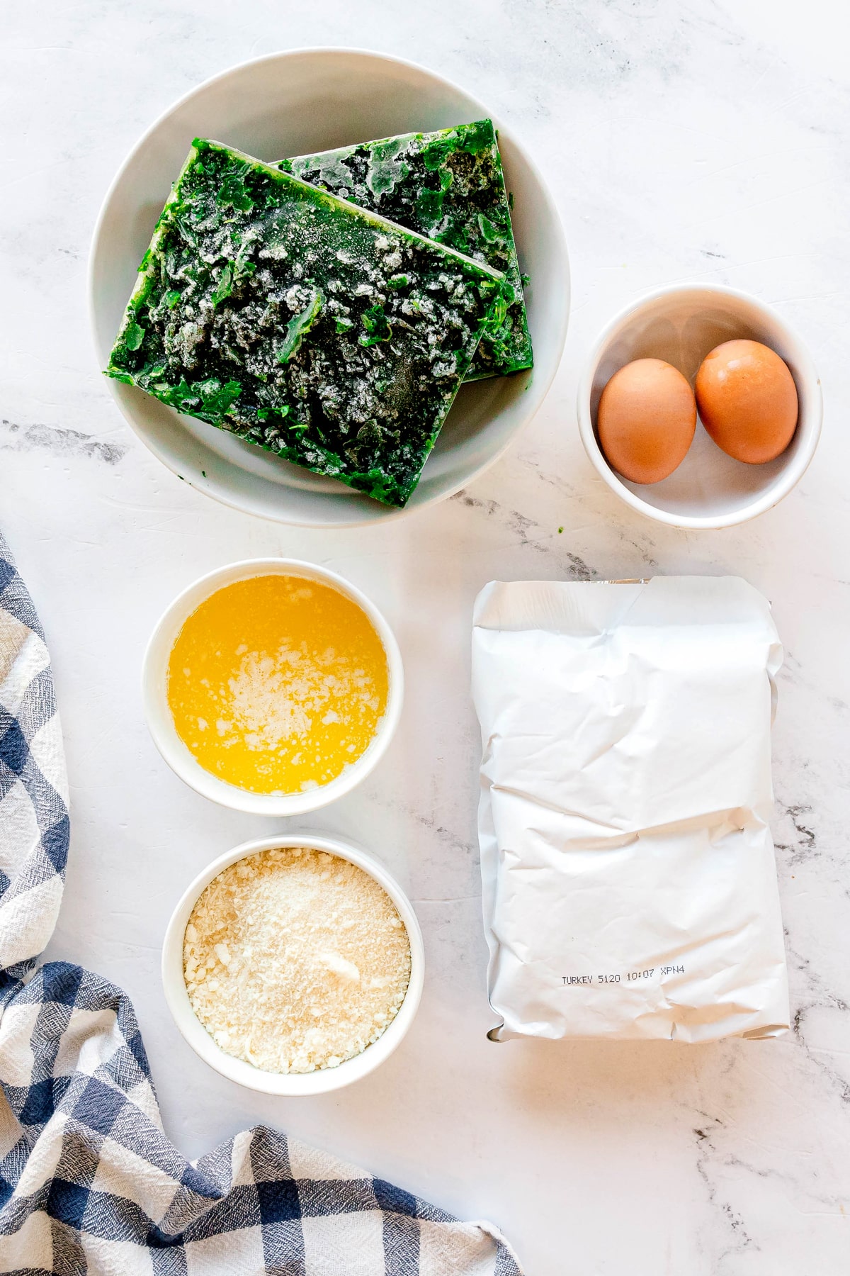 Ingredients for Spinach Balls include a bowl of frozen spinach, two eggs, melted butter, grated cheese, and stuffing mix on a marble surface with a blue checkered cloth.