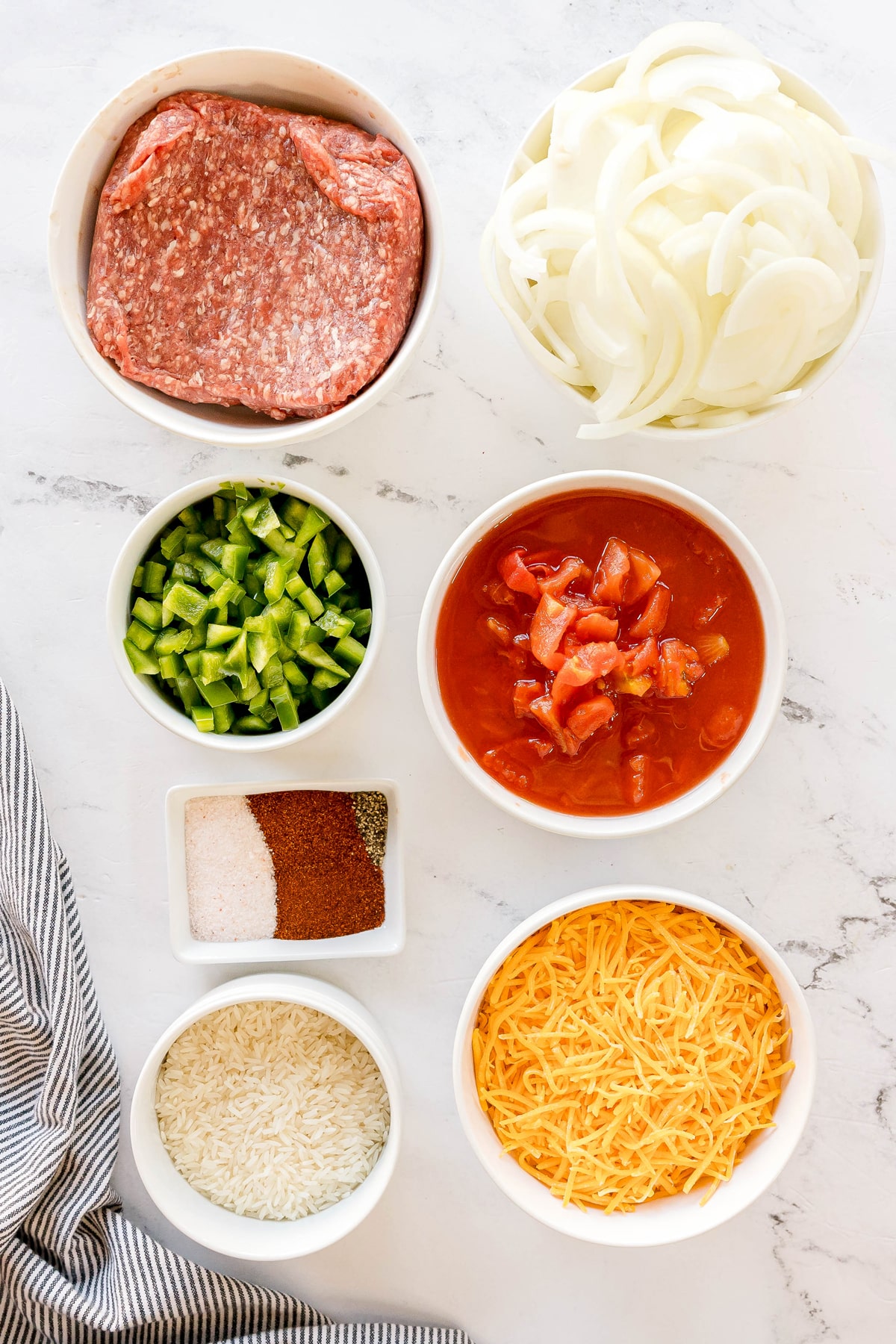 Bowls of ingredients for Texas Hash containing ground beef, sliced onions, chopped green bell peppers, diced tomatoes, shredded cheddar cheese, uncooked rice, salt, chili powder, and black pepper.