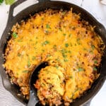 A cast iron skillet filled with Texas Hash casserole, topped with melted cheddar cheese, with a serving spoon scooping out a hearty portion.