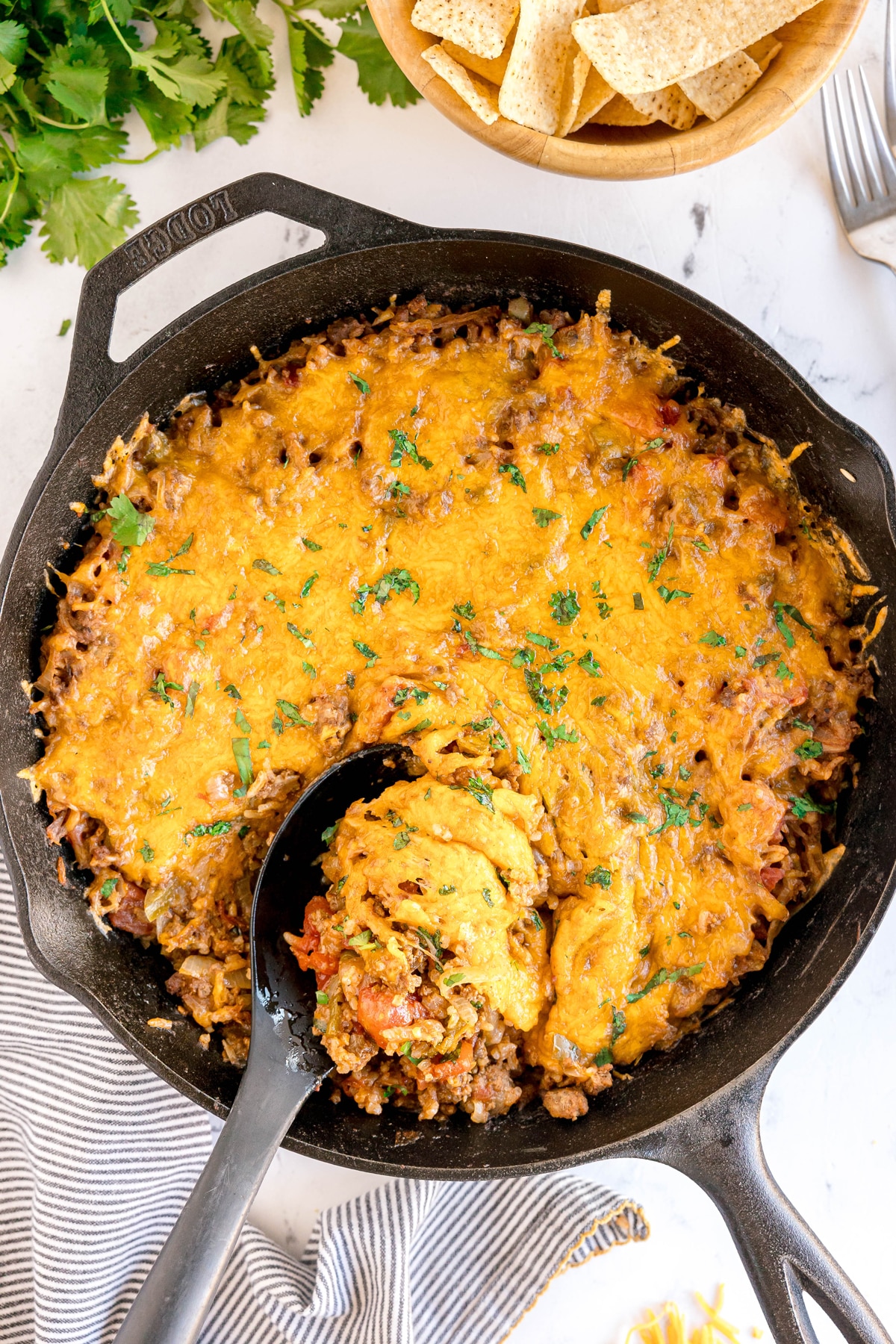 A cast iron skillet filled with baked cheesy Texas Hash casserole, containing ground meat and topped with melted cheese, with a serving spoon scooping a portion. Tortilla chips and cilantro are nearby.