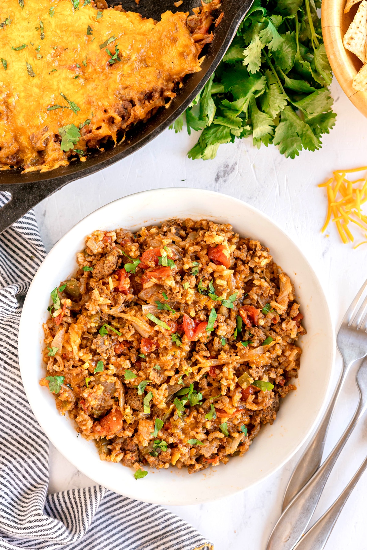 A white bowl filled with a Texas Hash including ground beef, rice, tomato, and cheese mixture sits on a table near a skillet of baked casserole and fresh cilantro.