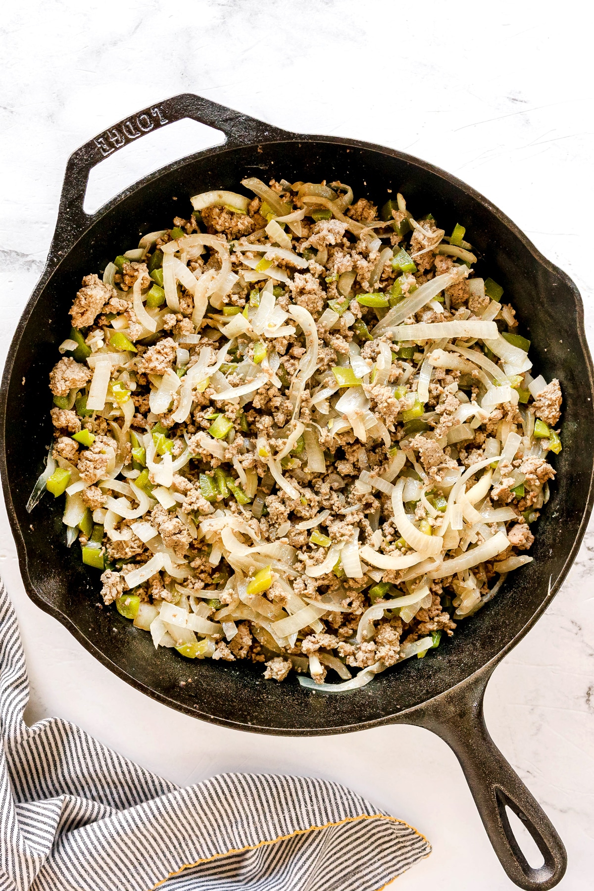 A cast iron skillet filled with cooked ground meat, sliced onions, and chopped green bell peppers on a white surface for Texas Hash recipe.