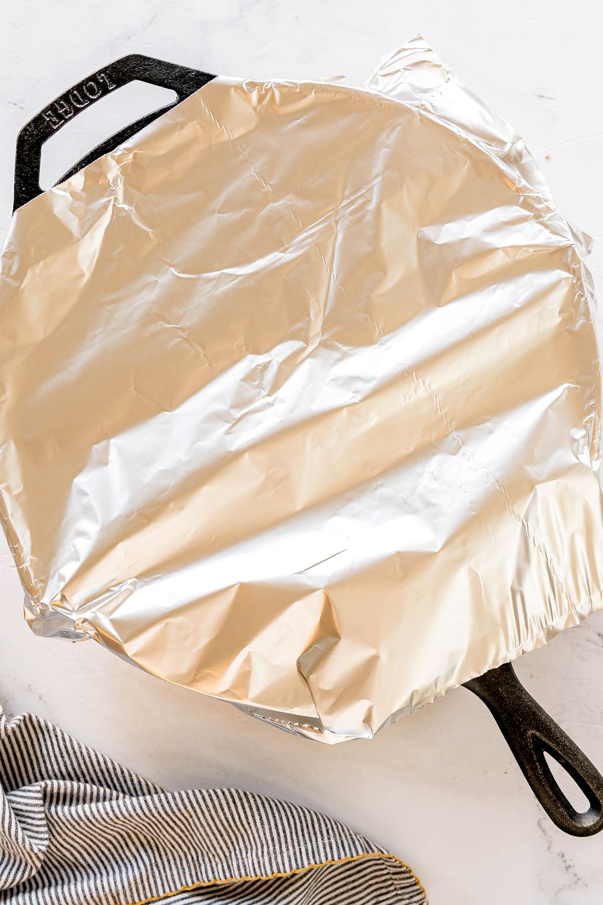 A black cast iron skillet covered with a sheet of aluminum foil, perfect for baking Texas Hash, sits beside a striped kitchen towel on a white surface.