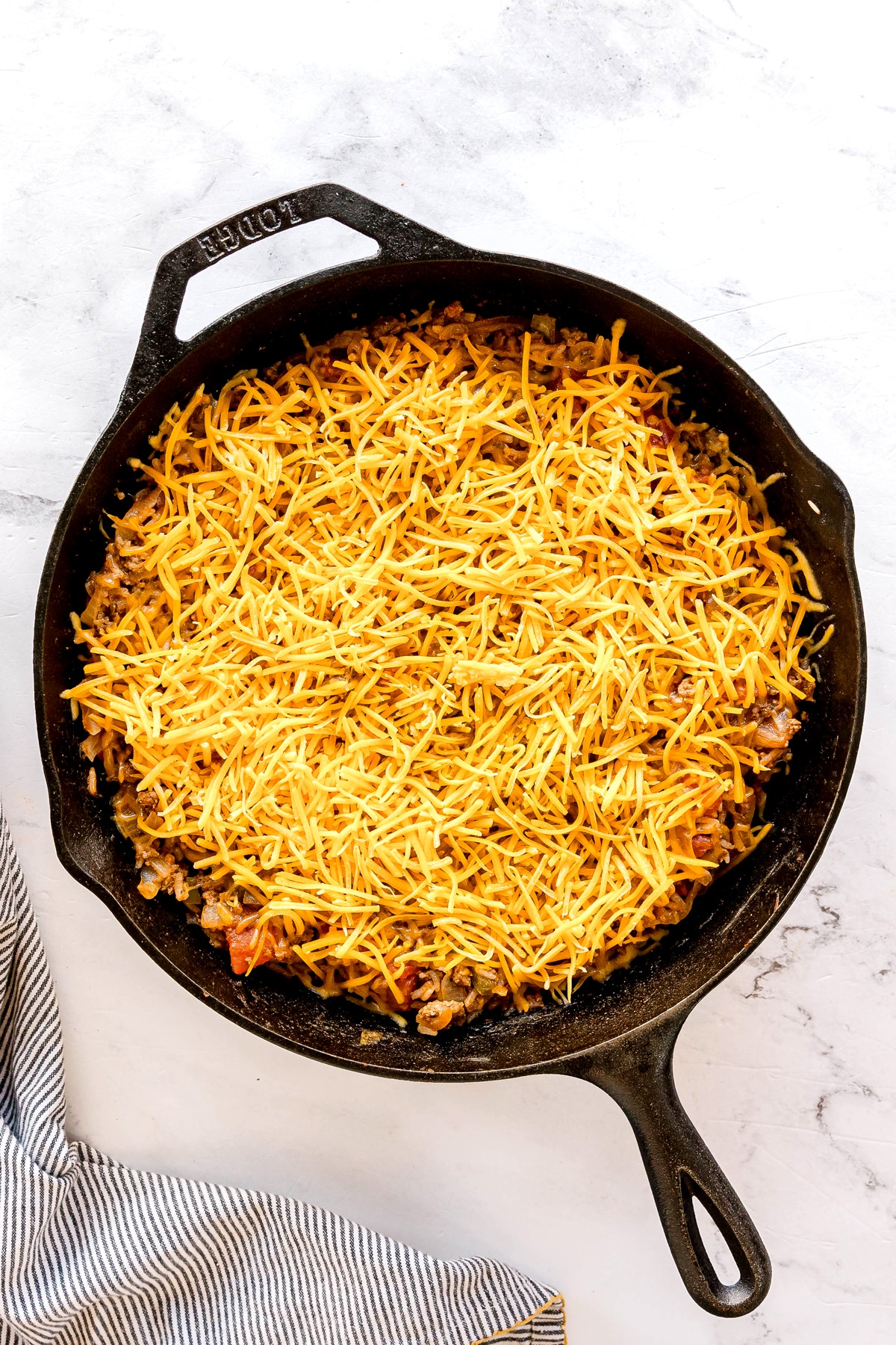 Adding shredded cheese on black cast iron skillet for Texas Hash recipe, placed on a white surface with a striped cloth nearby.