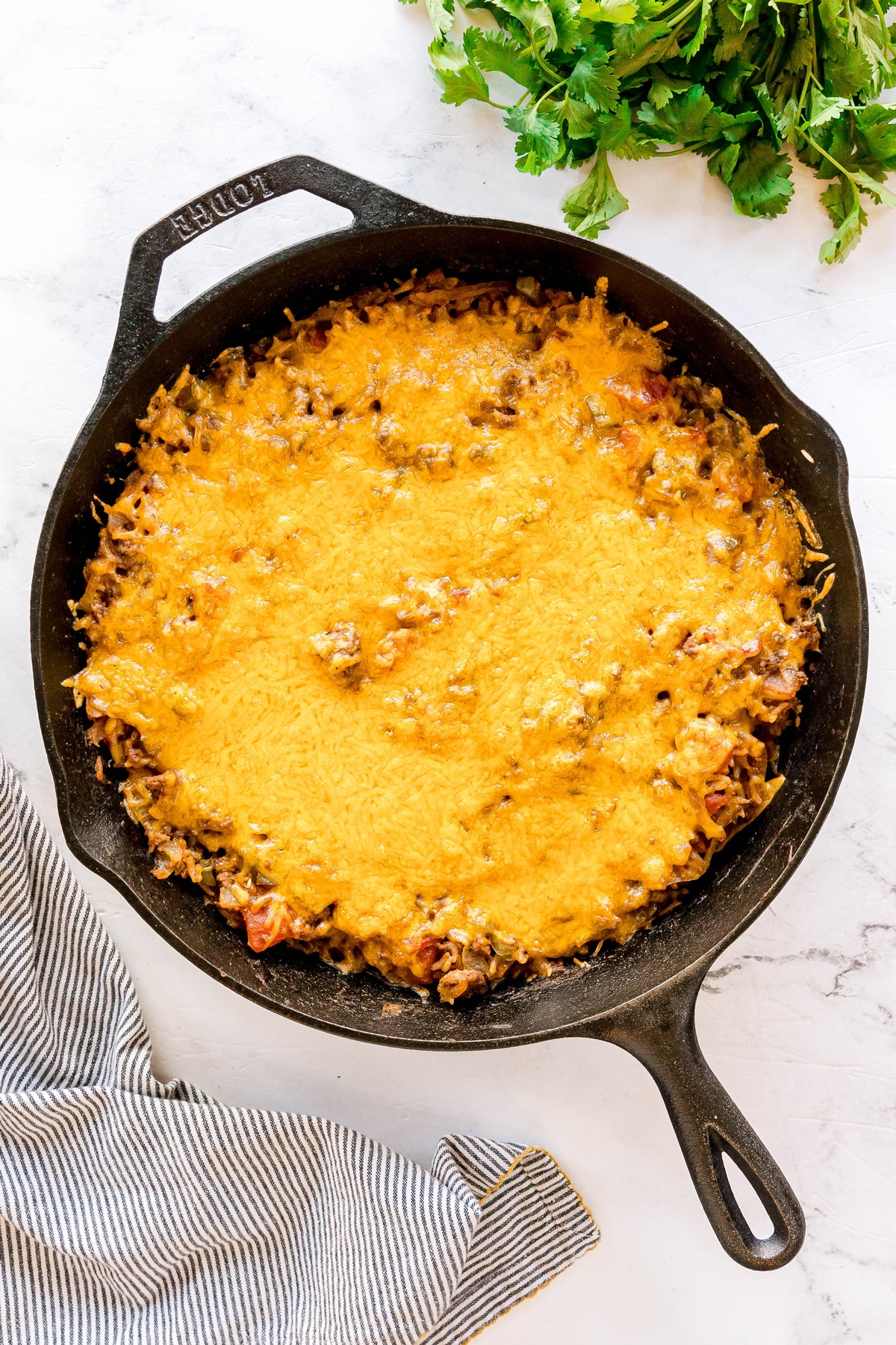 A cast iron skillet filled with a baked cheesy Texas Hash casserole sits on a white surface, with fresh cilantro and a striped cloth nearby.