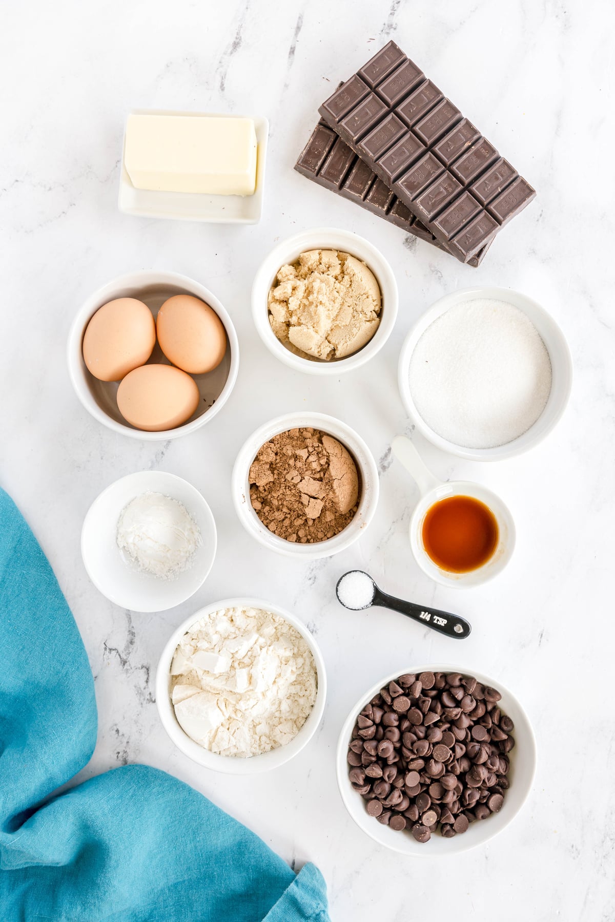 Overhead view of baking ingredients for Easy Brownie Recipe: unsalted butter, semi-sweet chocolate bars, eggs, granulated sugar, brown sugar, cocoa powder, vanilla extract, all-purpose flour, cornstarch, salt, and semi-sweet chocolate chips on a marble surface.