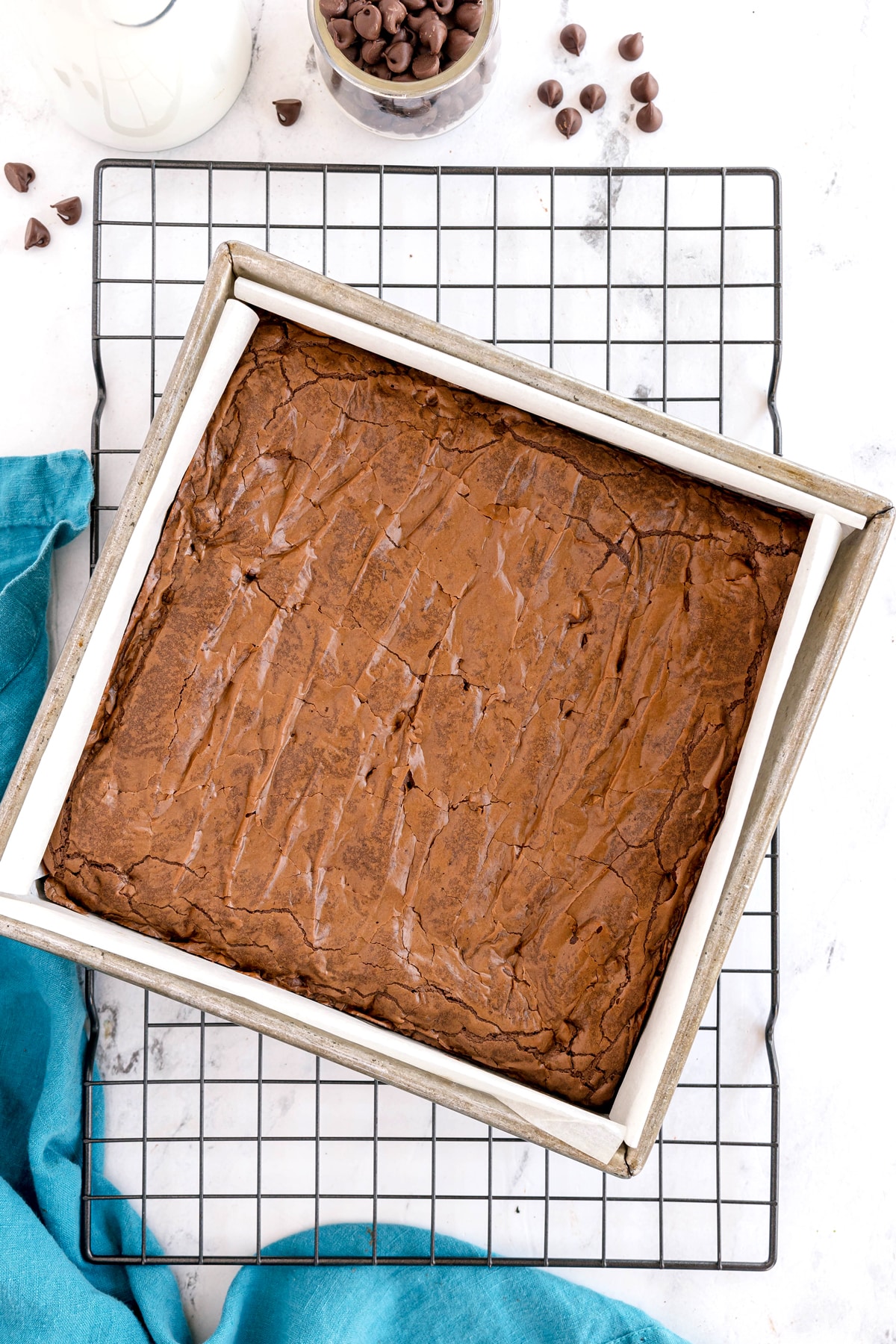 A pan of freshly baked brownies on a cooling rack, with parchment paper lining the pan and a few chocolate chips and a glass of milk nearby.