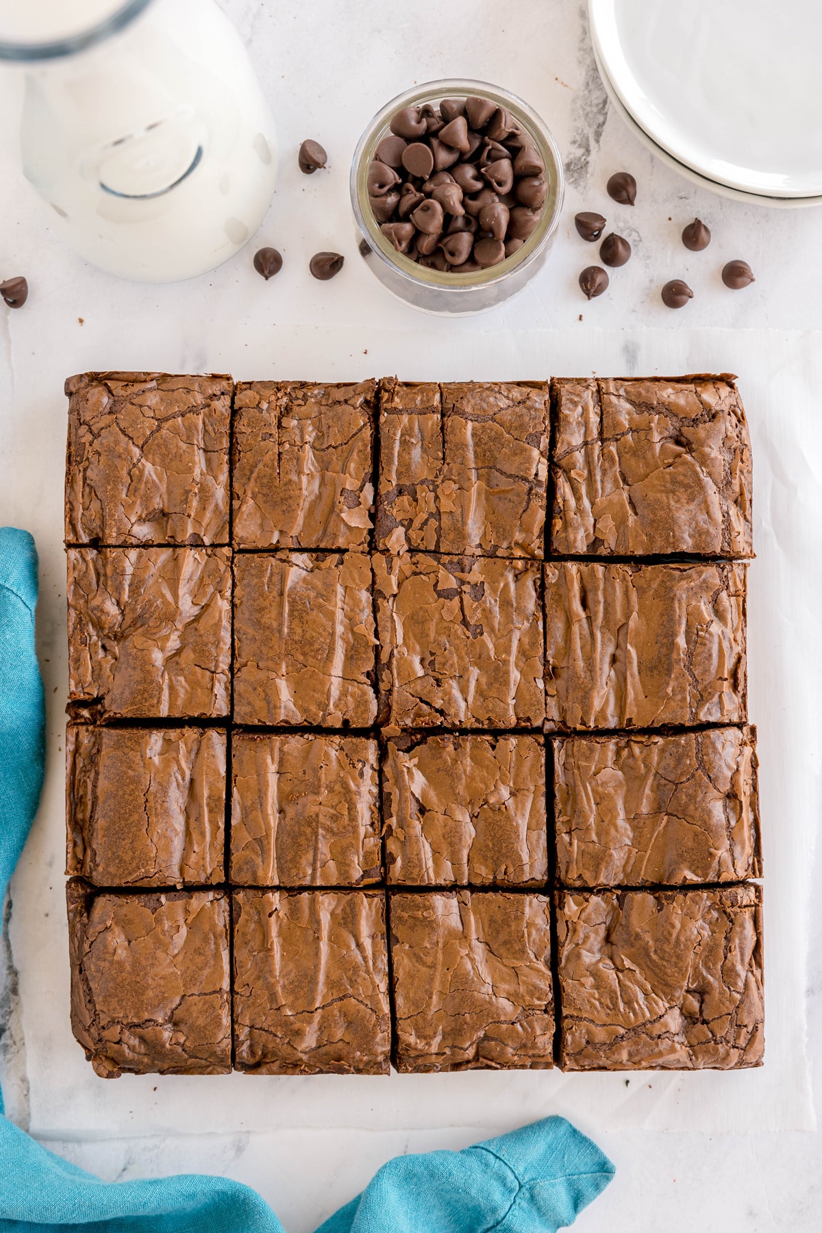 A batch of 16 square-cut brownies made from an easy brownie recipe on parchment paper, next to a jar of chocolate chips, a bottle of milk, and a blue cloth on a marble surface.