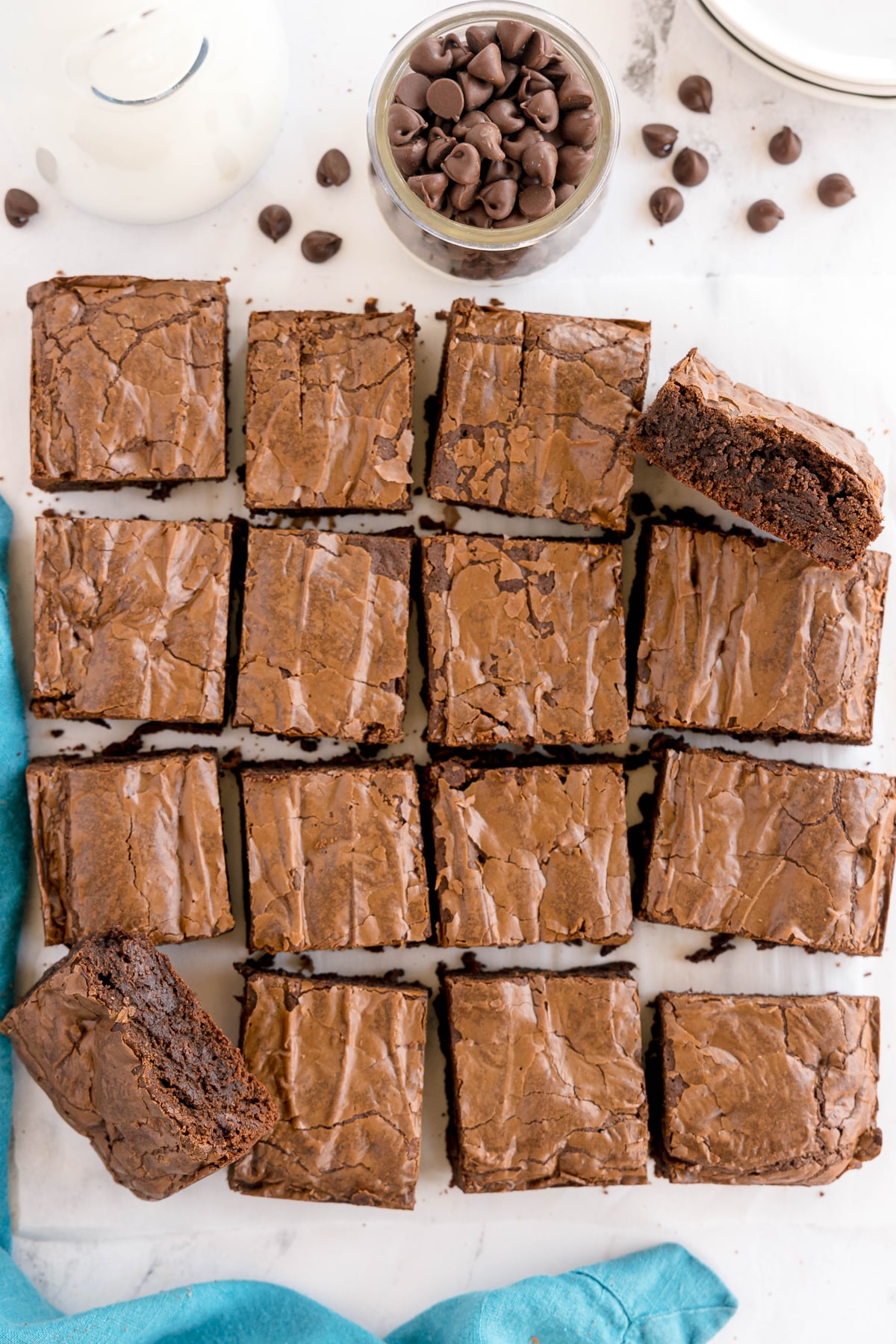 A batch of 16 chocolate brownies on parchment paper, with a glass of chocolate chips and a bottle of milk nearby.