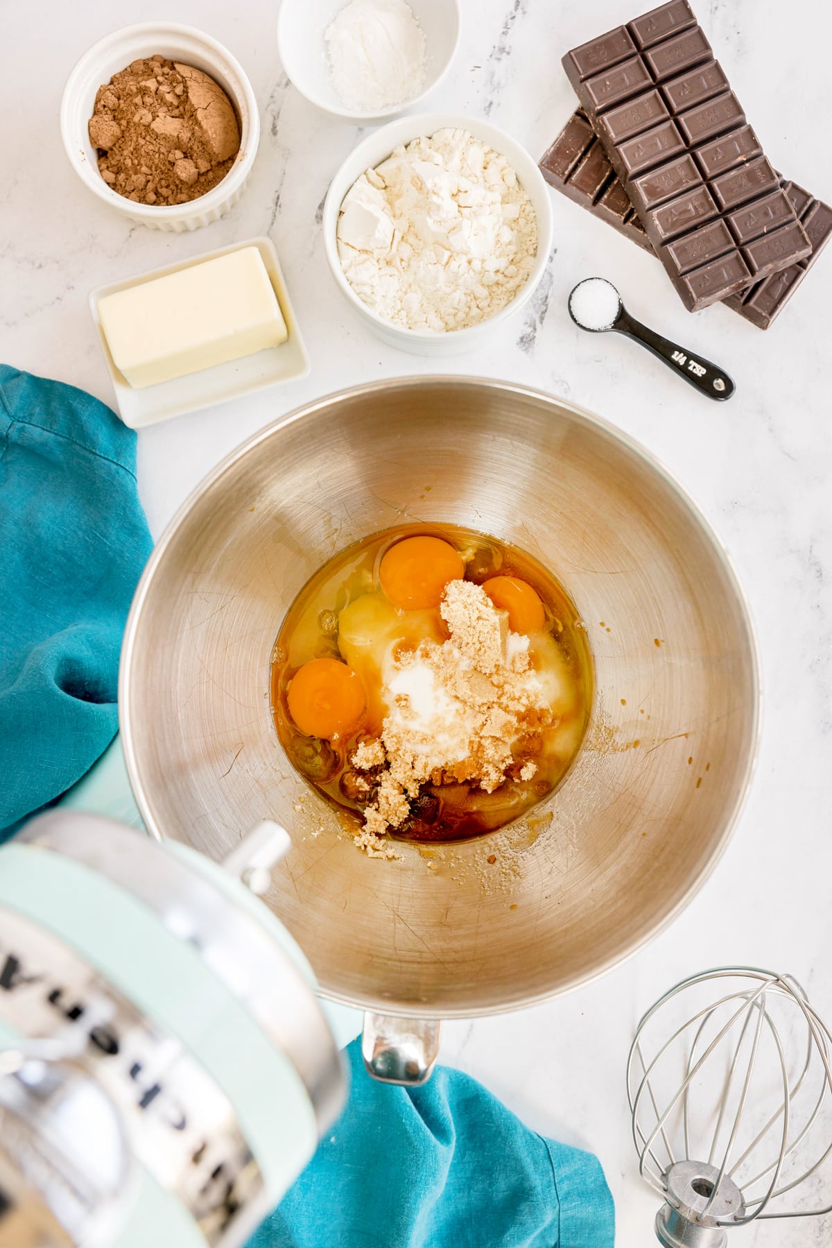 First step in preparing Easy Brownie Recipe is to use a mixing bowl with eggs, sugar, and vanilla is on a counter surrounded by butter, cocoa powder, flour, chocolate bars, a measuring spoon, and a stand mixer.
