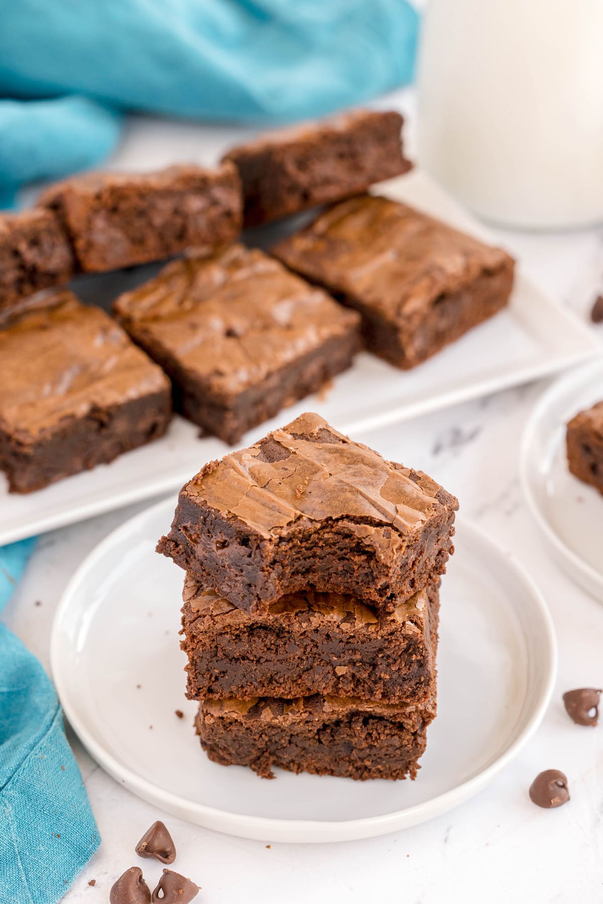 A stack of three chocolate brownies on a white plate, with more brownies on a tray and a glass of milk in the background.