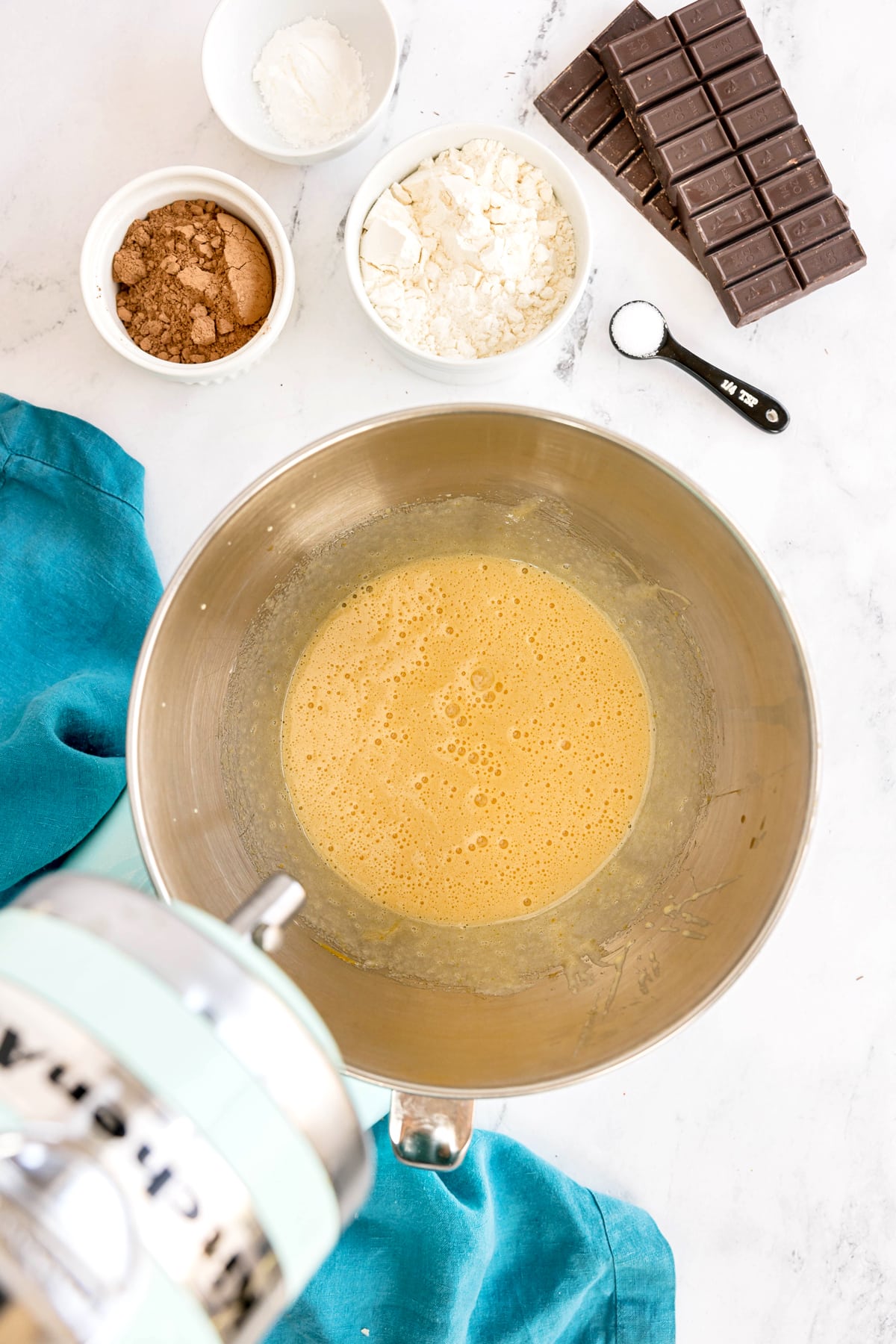 A mixing bowl with beaten eggs on a counter, surrounded by bowls of flour, cocoa powder, cornstarch, a chocolate bar, and a measuring spoon. This is one step in preparing Easy Brownie Recipe.