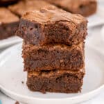 A stack of three chocolate brownies is placed on a white plate, with more brownies visible in the background.
