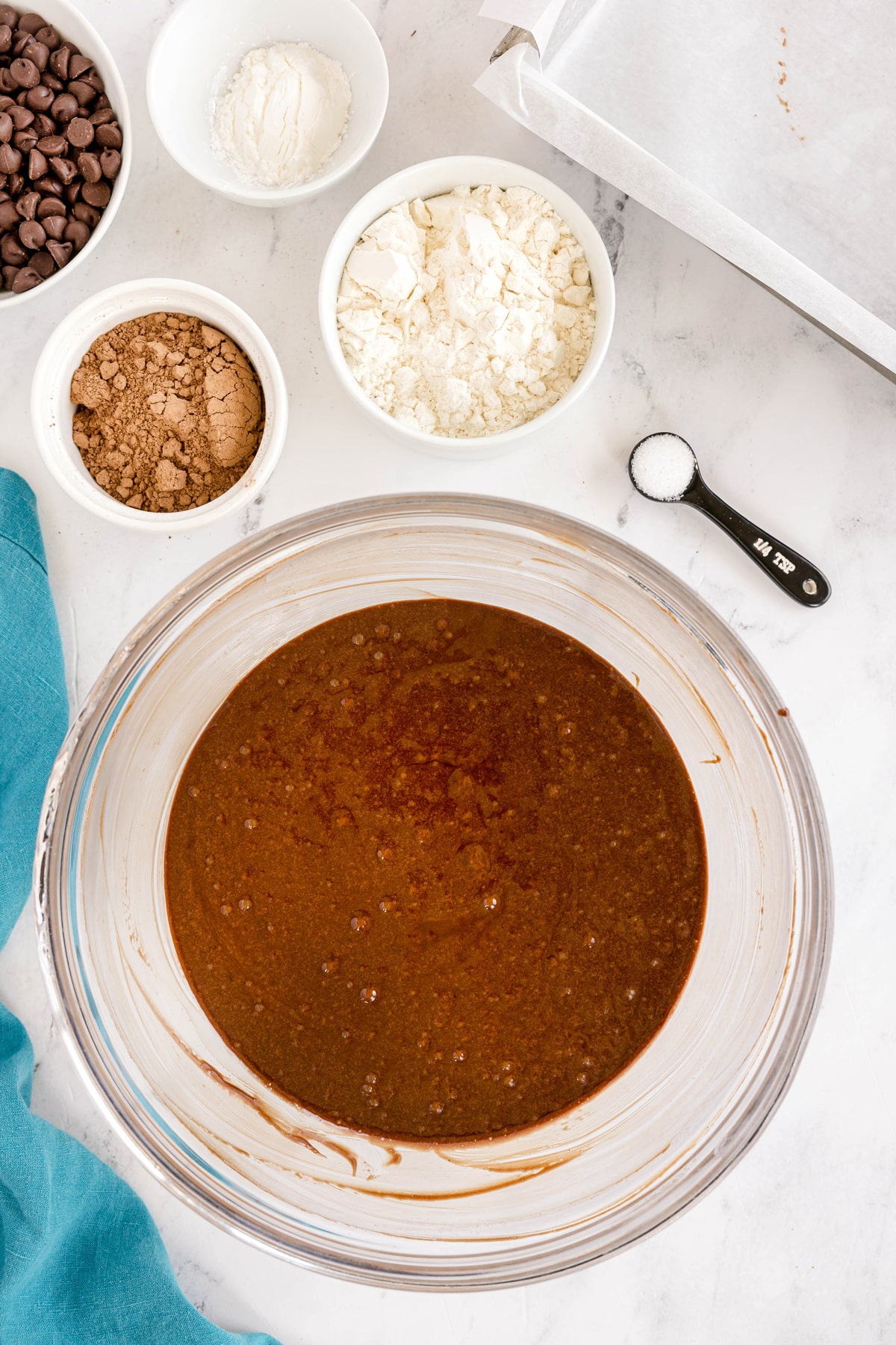 A glass bowl with Easy Brownie Recipe sits on a marble surface, surrounded by bowls of cocoa powder, flour, chocolate chips, and a measuring spoon of salt.