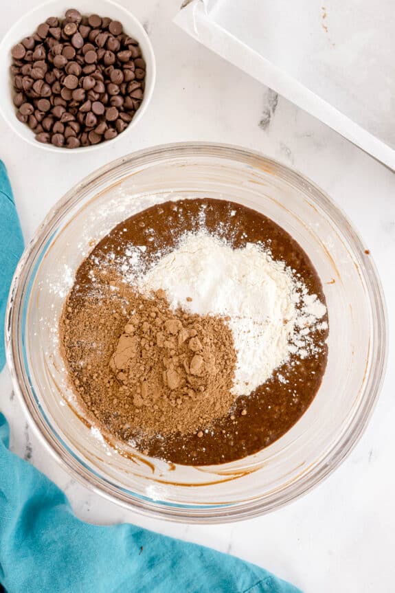 Next step in preparing Easy Brownie Recipe is to mix cocoa powder, flour, and wet ingredients, next to a bowl of chocolate chips and a blue cloth on a marble surface.