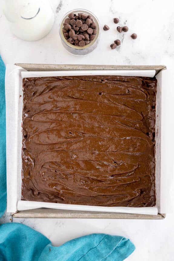A baking pan lined with parchment paper filled with brownie batter, next to a small bowl of chocolate chips and a bottle of milk on a marble surface.