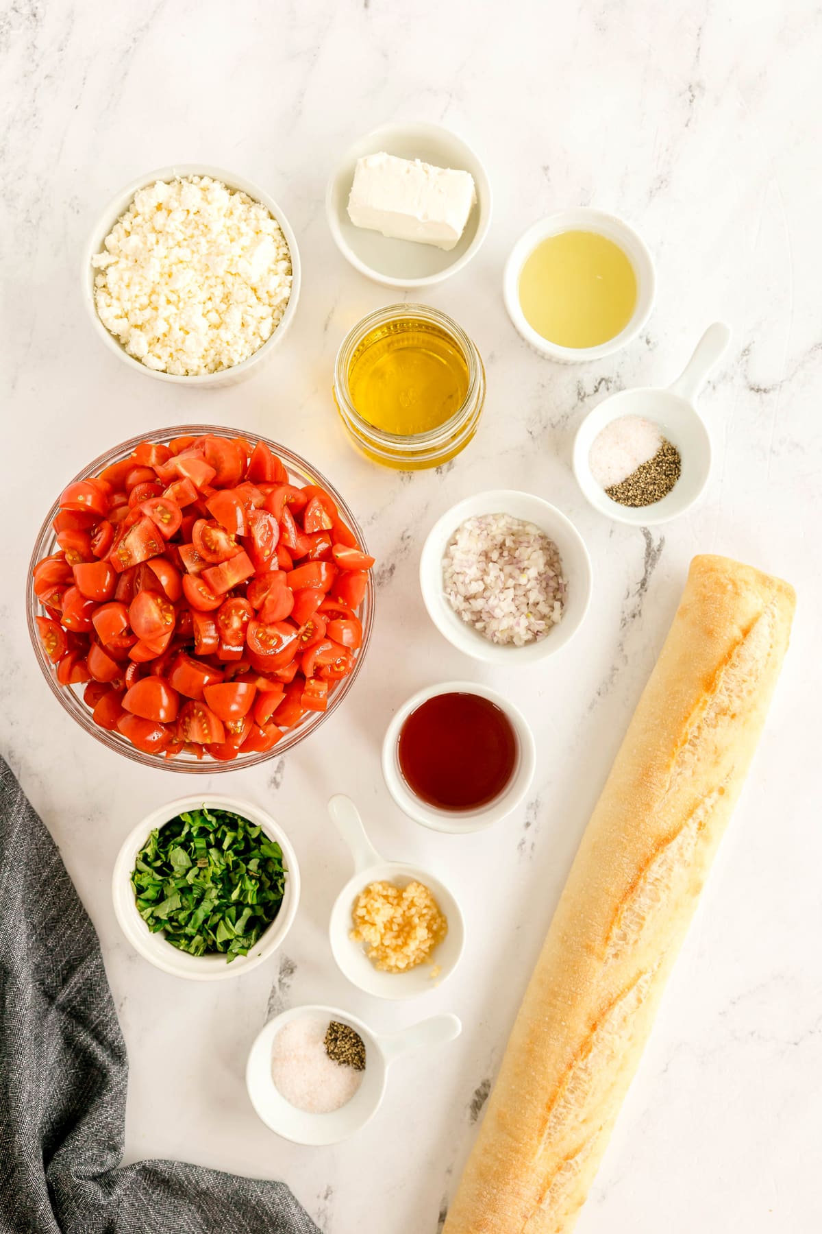 Ingredients for Whipped Feta Tomato Crostini including chopped tomatoes, baguette, olive oil, vinegar, feta cheese, garlic, herbs, shallots, salt, pepper, and cheese, arranged on a white surface.