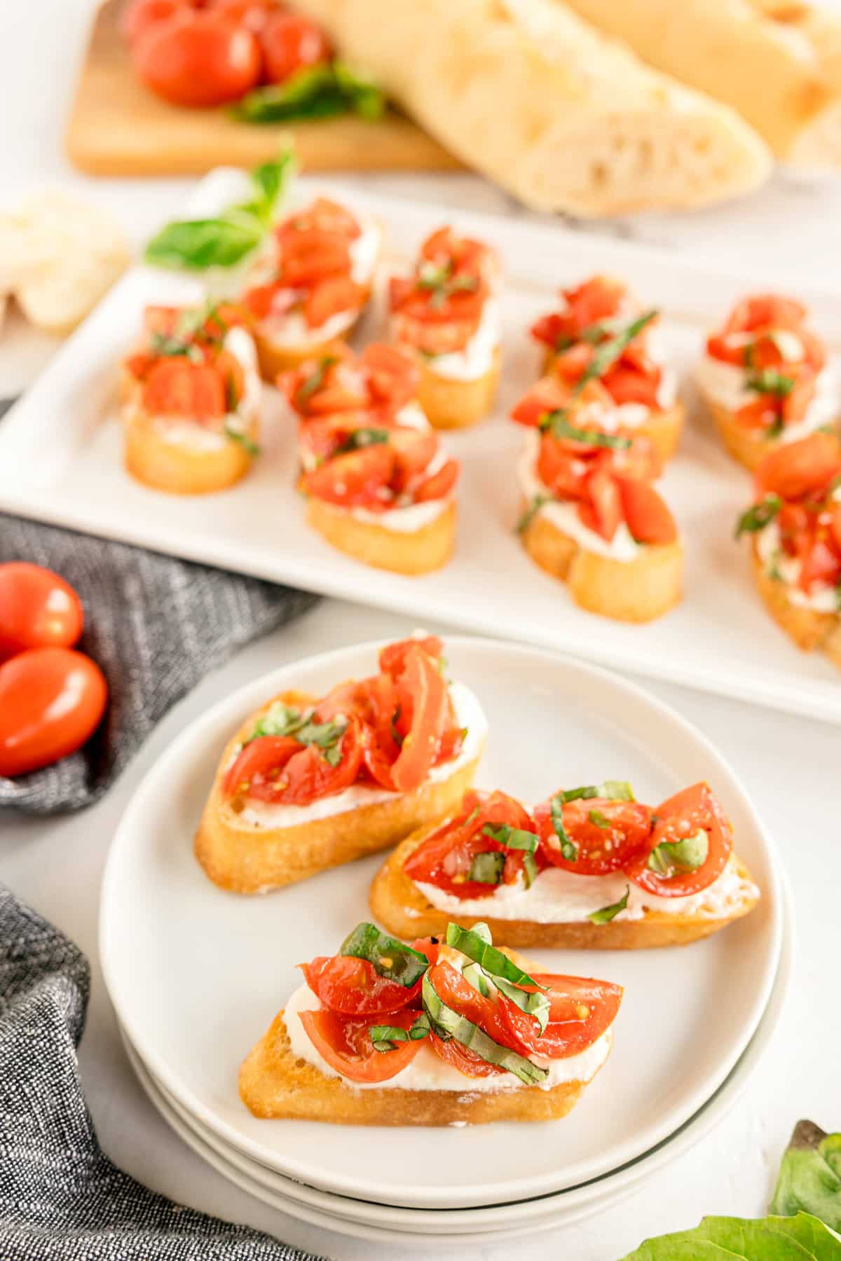 Slices of toasted bread topped with whipped feta crostini, chopped tomatoes, basil, and cheese are arranged on a plate and a serving tray, with fresh tomatoes and bread in the background.