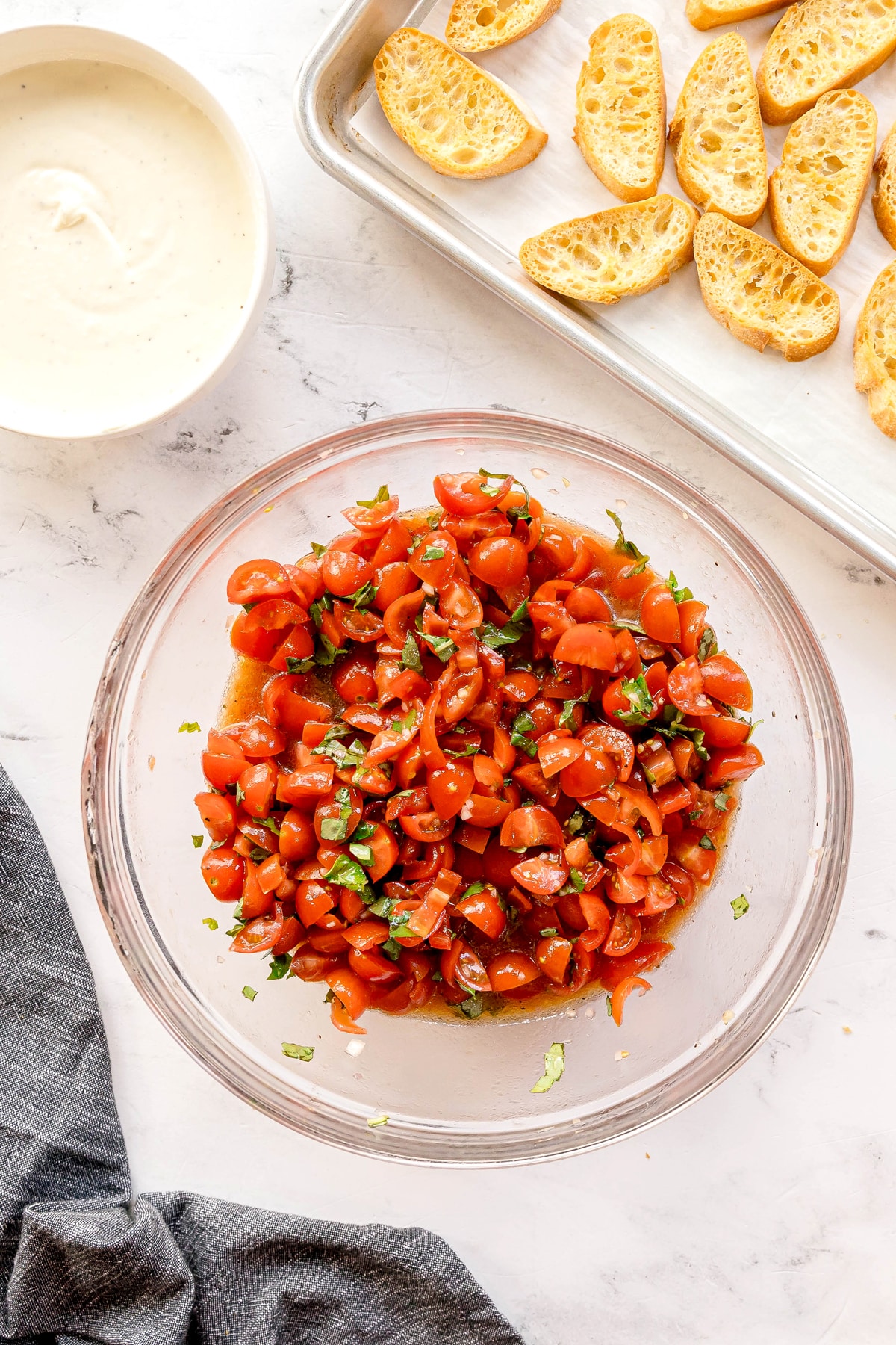 A glass bowl of Whipped Feta Tomato Crostini mixture with basil, sits on a marble surface next to a tray of toasted baguette slices and a small bowl of white cream cheese sauce.