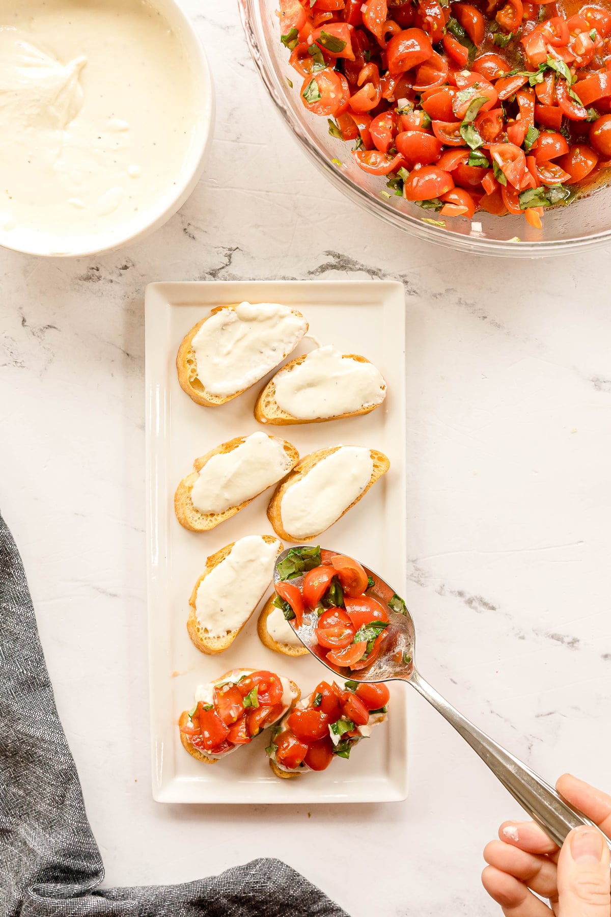 A hand spoons Whipped Feta Tomato Crostini mixture onto toasted bread slices topped with melted cheese, next to a bowl of the mixture and a bowl of white sauce on a white surface.