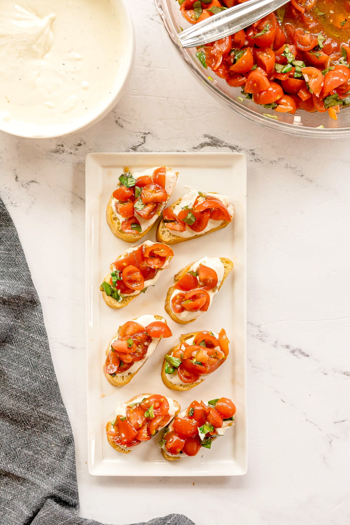 Whipped Feta Tomato Crostini on rectangular white plate with seven toasted baguette slices topped with diced tomatoes and herbs, next to a bowl of tomato mixture and a bowl of creamy dip on a white surface.