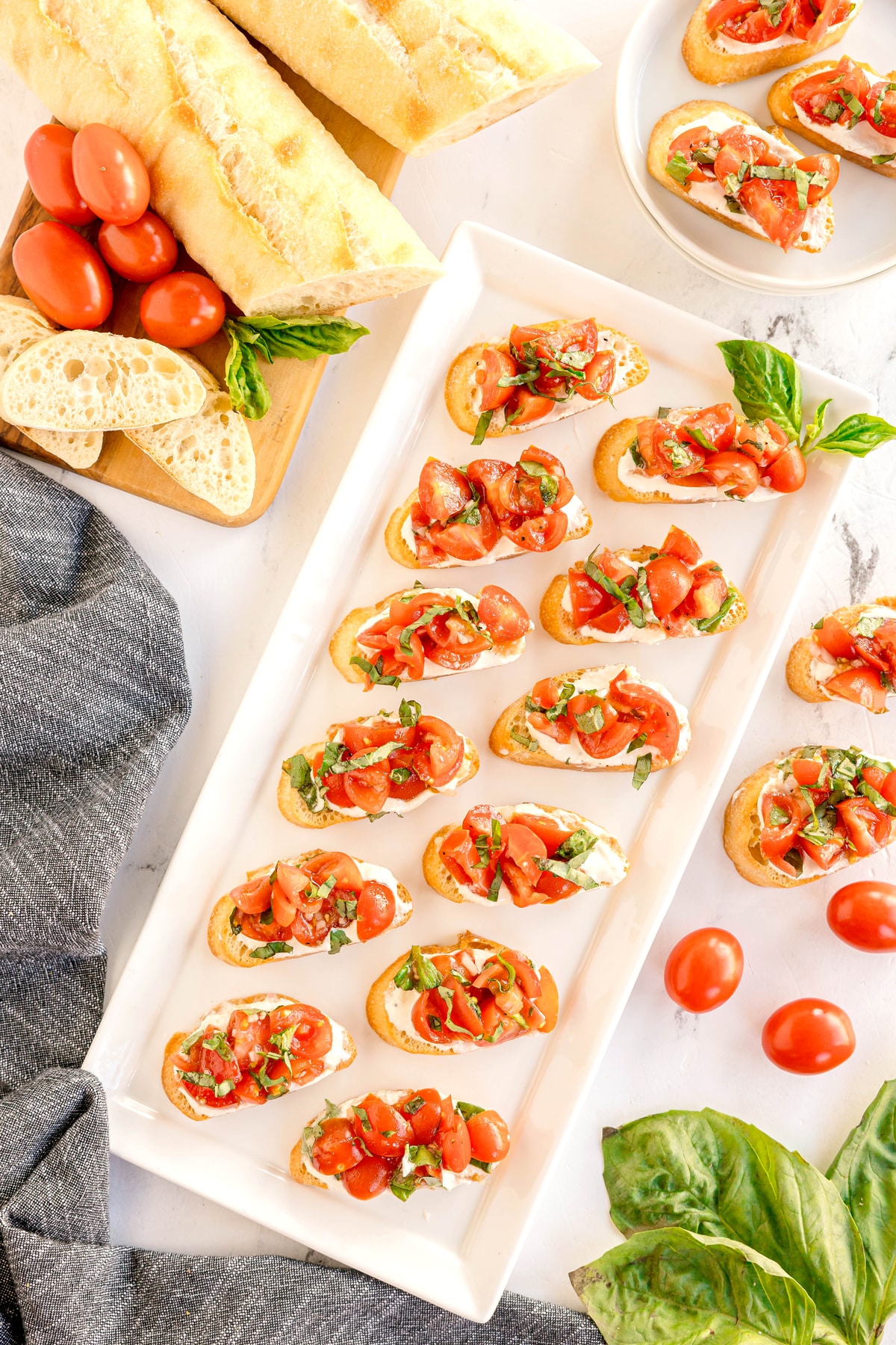 A platter of Whipped Feta Tomato Crostini topped with diced tomatoes and basil is displayed with fresh baguette slices, cherry tomatoes, and basil leaves on a white surface.
