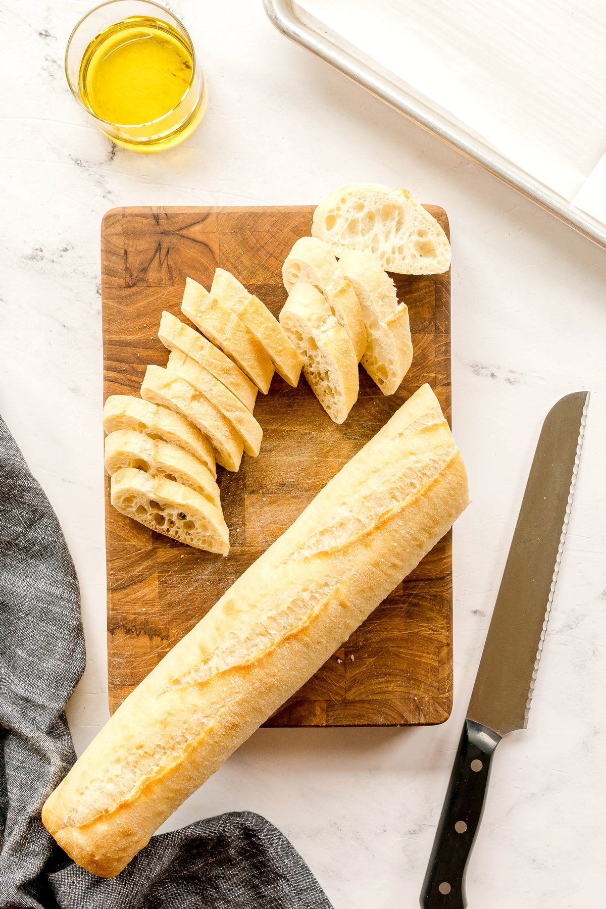 First step in making Whipped Feta Tomato Crostini is to slice baguette on a wooden cutting board with a bread knife. A glass of olive oil, and a gray cloth on a marble surface can also be seen.